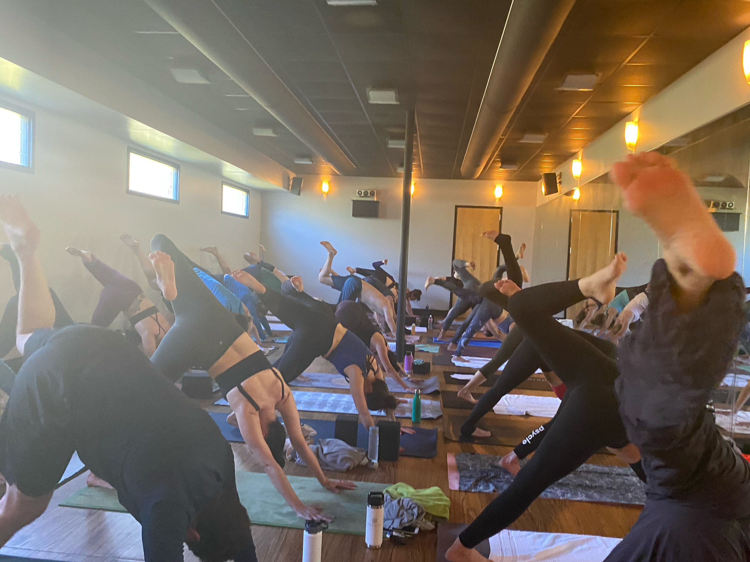 Group of people participating in a yoga class, practicing poses on yoga mats in a room with wooden floors and large windows.