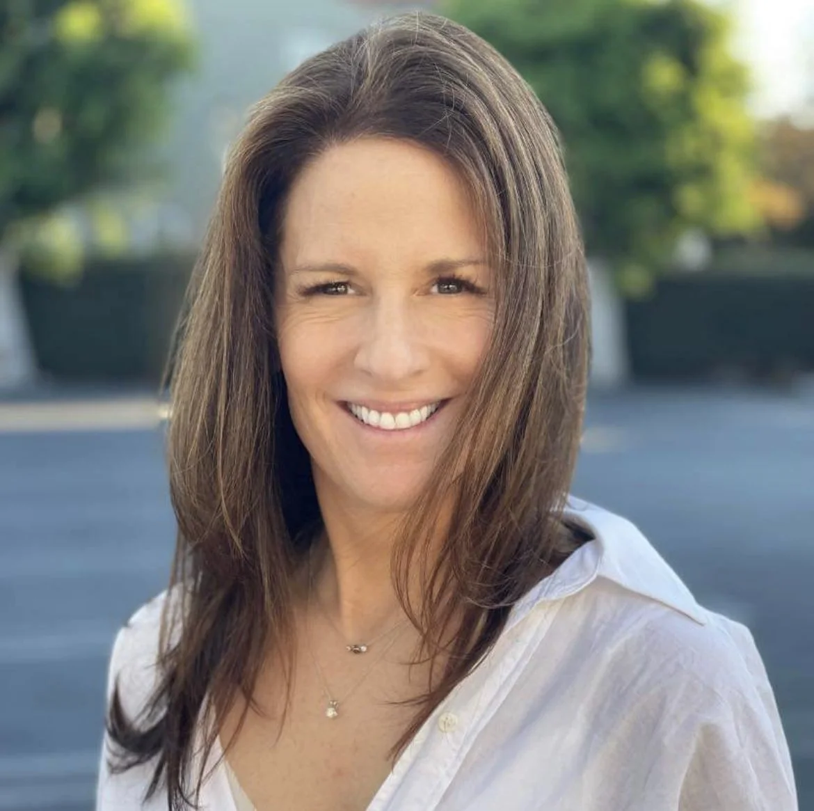 A woman with shoulder-length brown hair, smiling, wearing a white blouse, outdoors with blurred greenery and parking lot in the background.