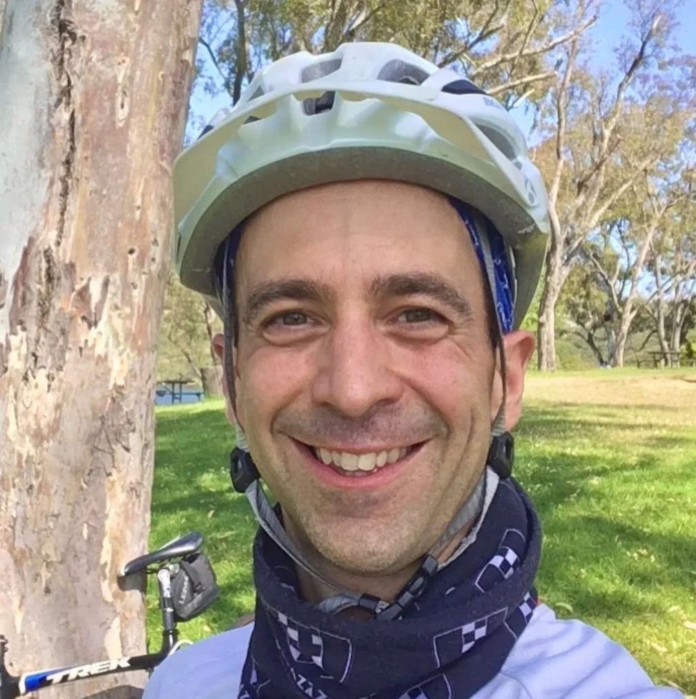 A man wearing a white bicycle helmet and a black neck gaiter, smiling outdoors near a tree, with a bicycle in the background.