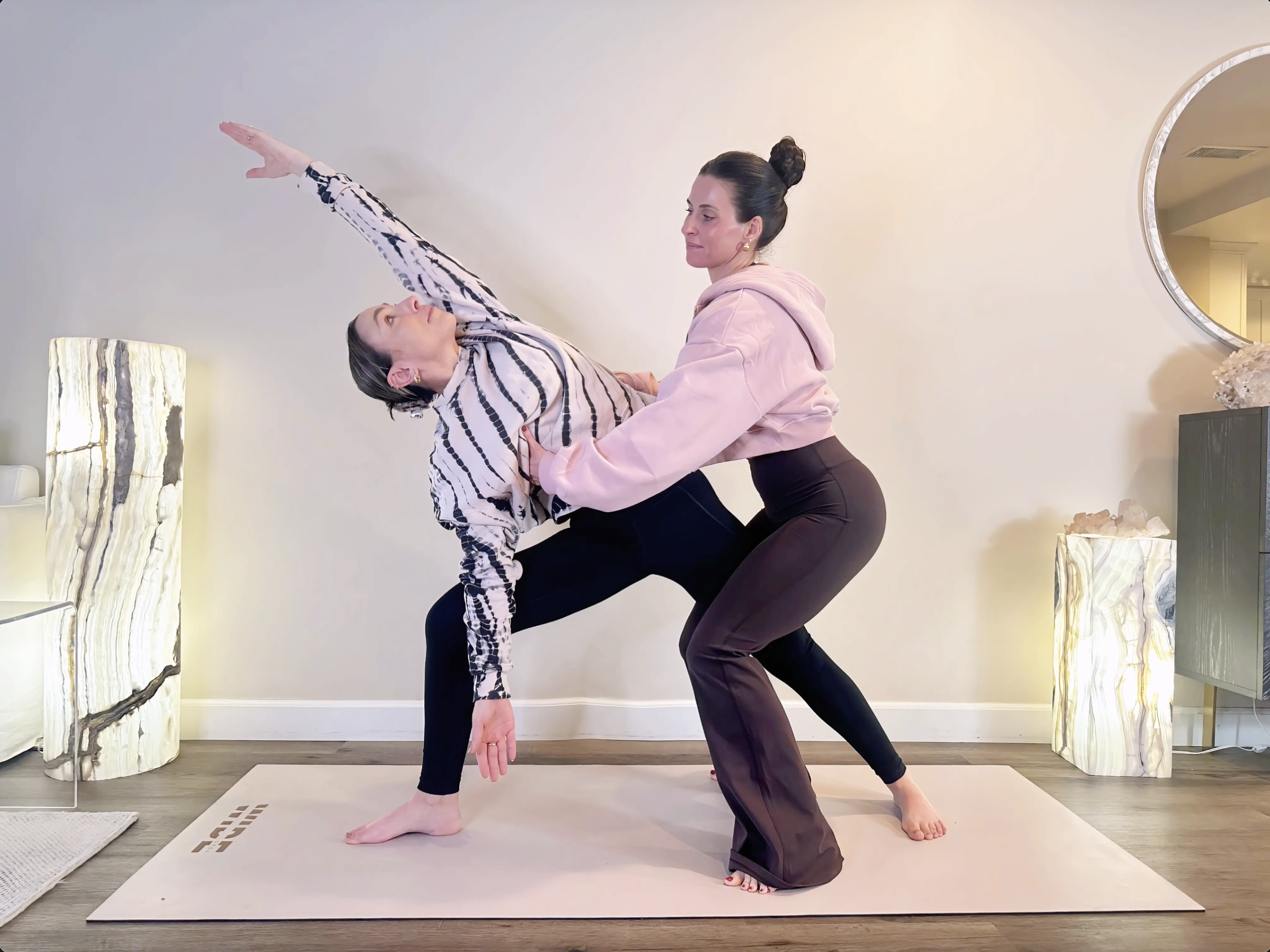 A woman sitting cross-legged on a mat by a swimming pool in a desert landscape with cacti and hills in the background during sunset.