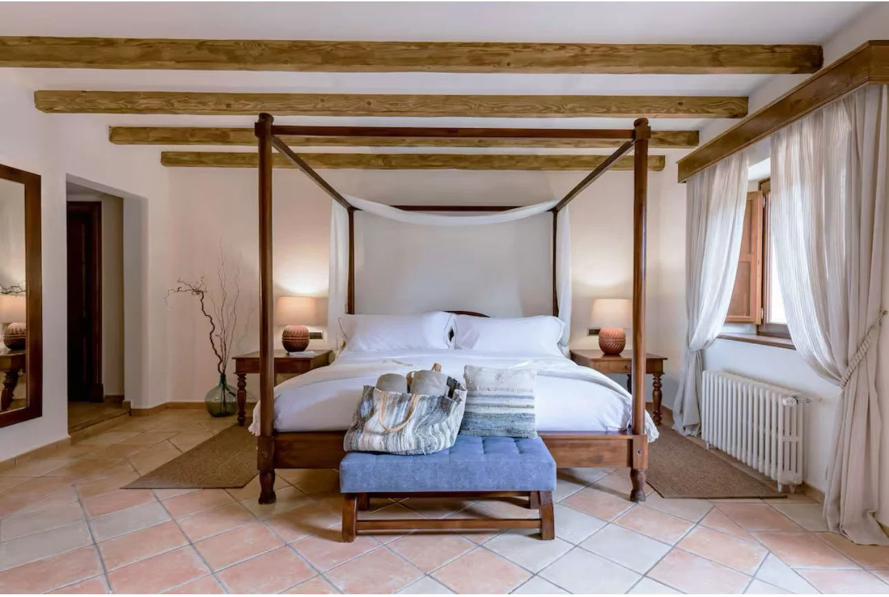 A bedroom with a four-poster bed, white bedding, and two bedside lamps, with curtains, a window, and tiled floor.