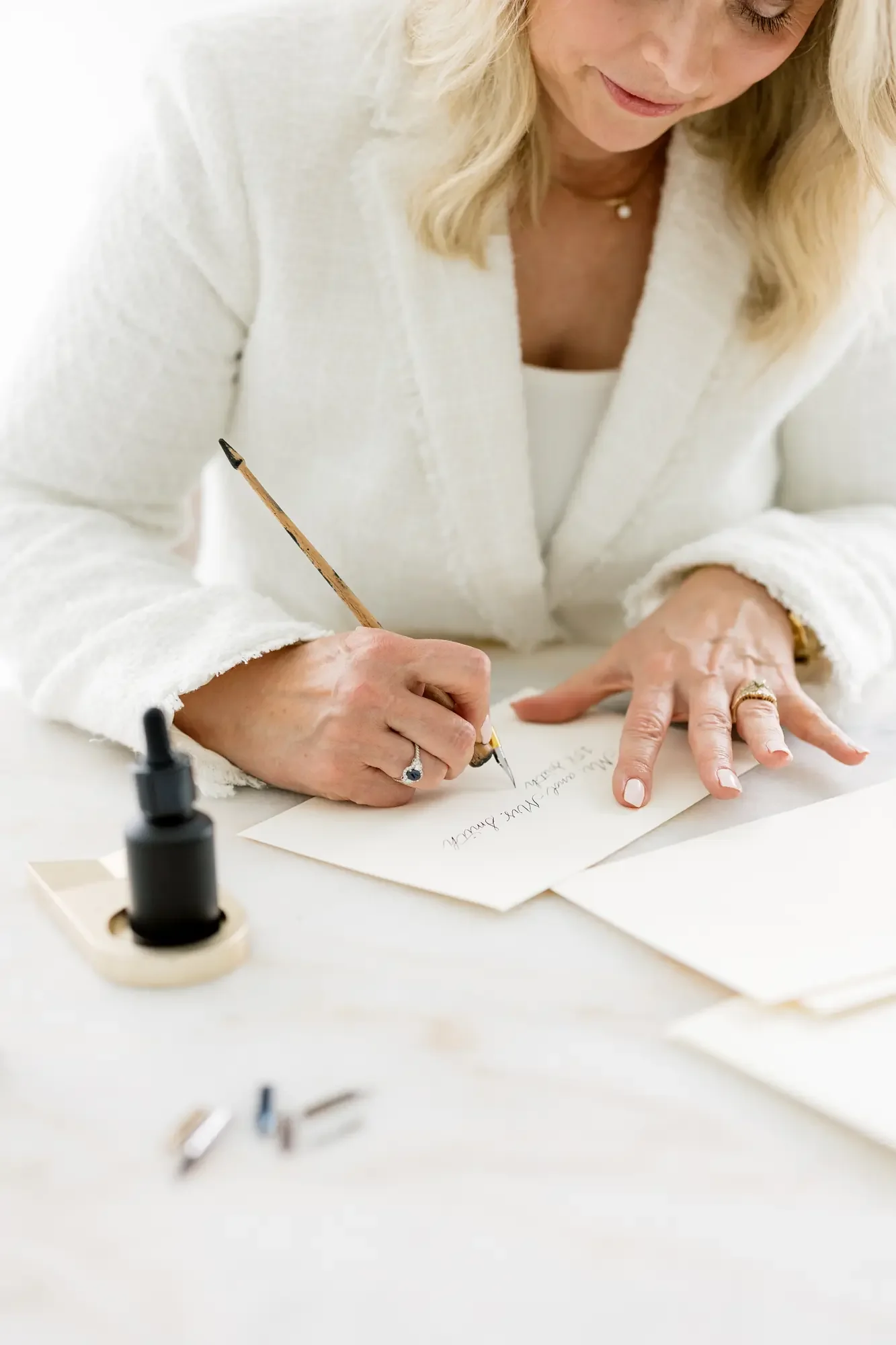 A woman with blonde hair, wearing a white tweed blazer, writes on a piece of paper with a calligraphy pen, sitting at a white desk with ink and paper.