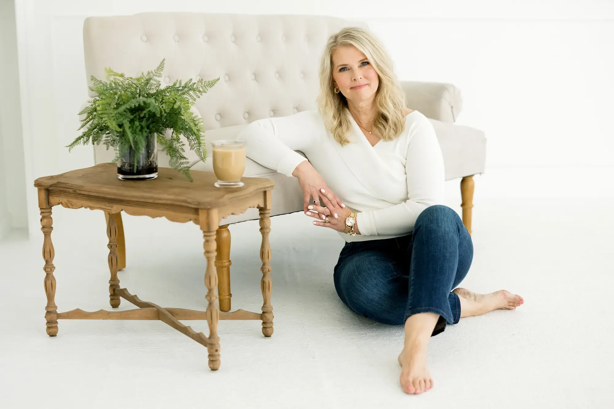 A woman with blonde hair and blue eyes, wearing a white long-sleeve top and blue jeans, sitting on the floor beside a wooden side table. The table has a potted green fern and a glass of coffee or tea. The background is mostly white with natural light illuminating the scene.