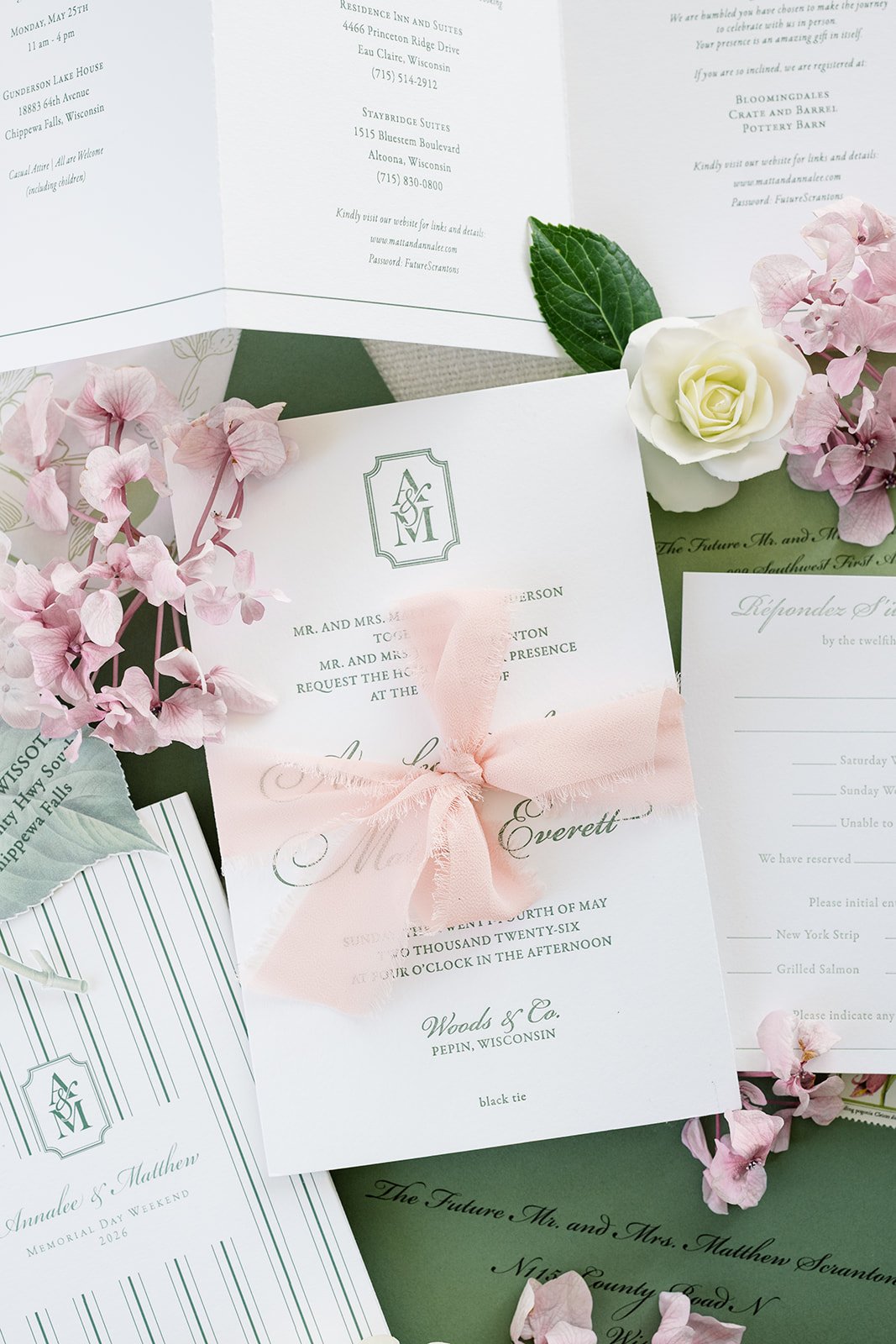 Letterpressed wedding invitation cards surrounded by pink and white flowers and green leaves, with a pink ribbon tied on invitation.