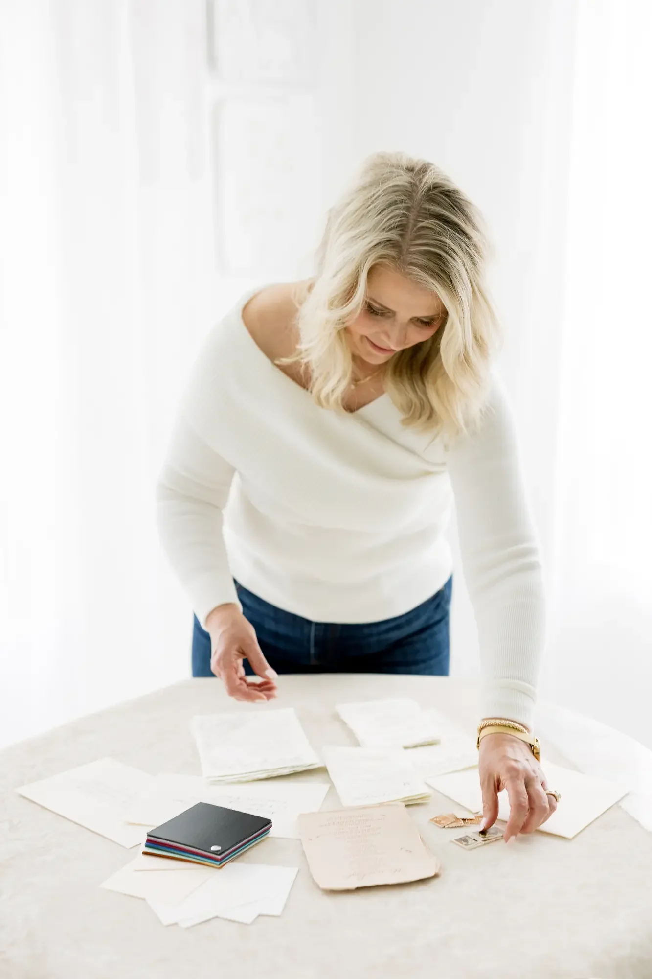 Woman with blonde hair in a white sweater standing at a table with papers and notebooks, examining photographs or documents.