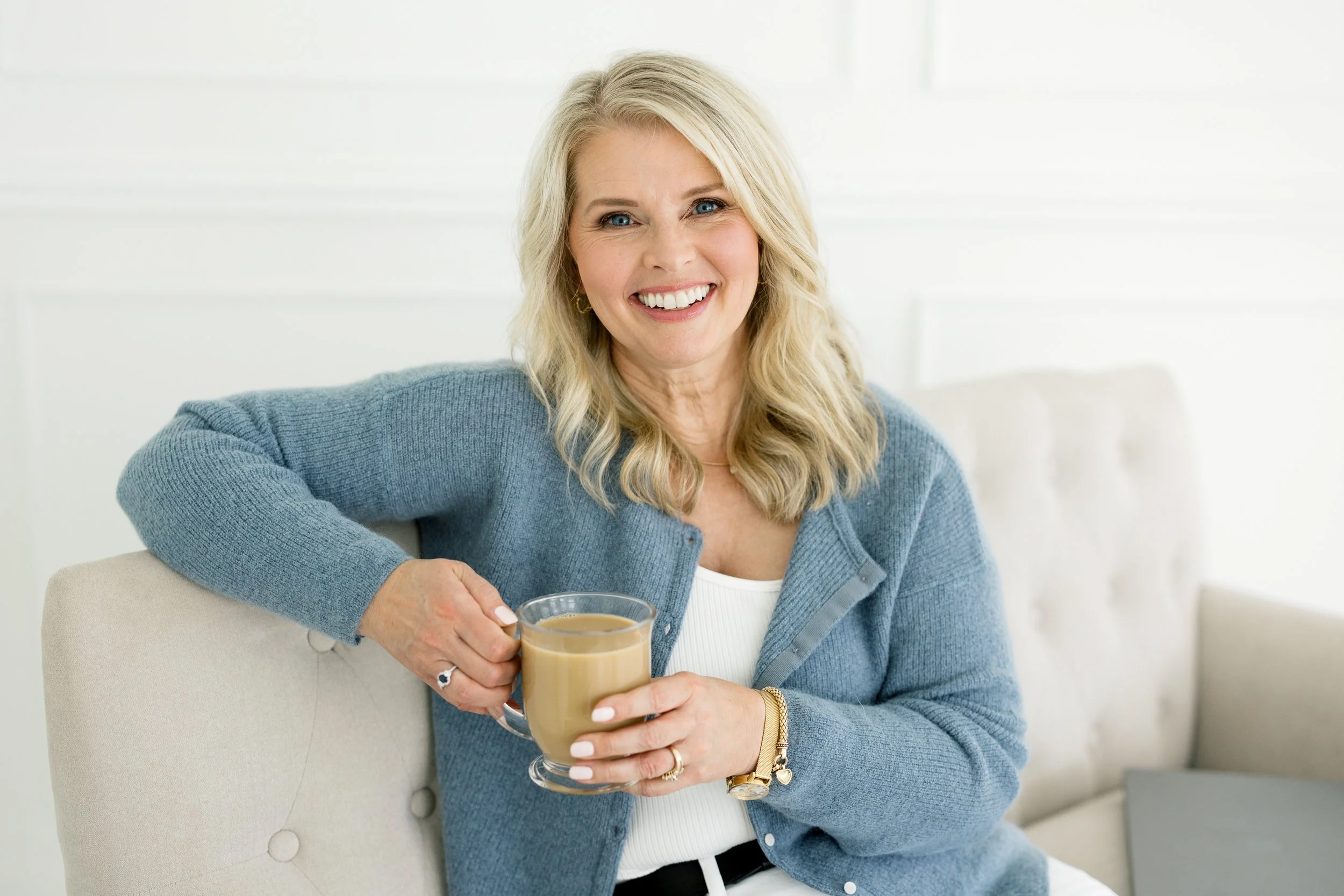 A smiling woman with blonde hair is sitting on a beige chair, holding a glass of coffee or tea. She is wearing a light blue cardigan, a white top, and gold jewelry, with a white background behind her.