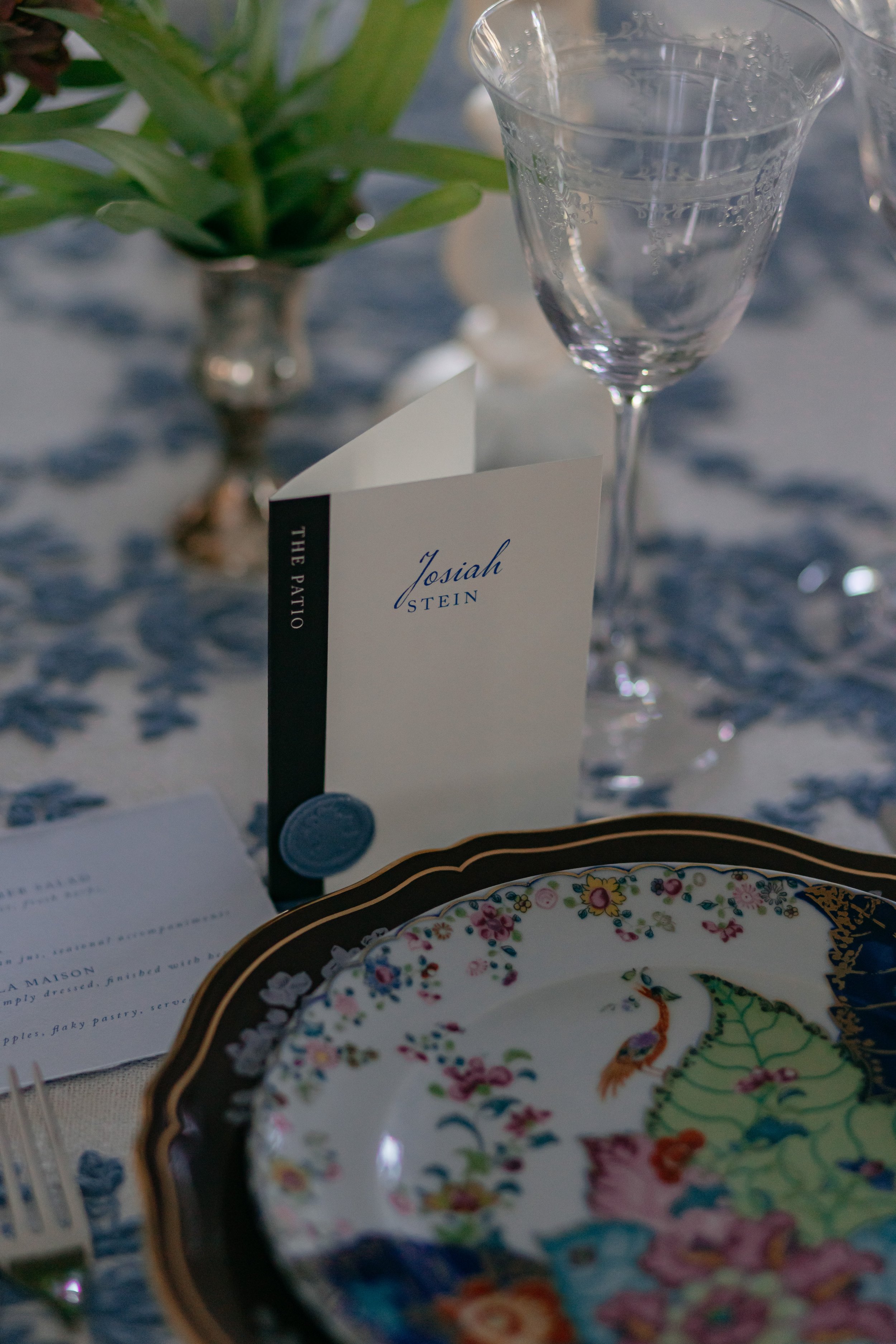 A wedding reception table setting with a vase of green leaves, a glass of water, a book-inspired place card with the name 'Josiah Stein',  a menu, and a decorative plate with an intricate, colorful design.