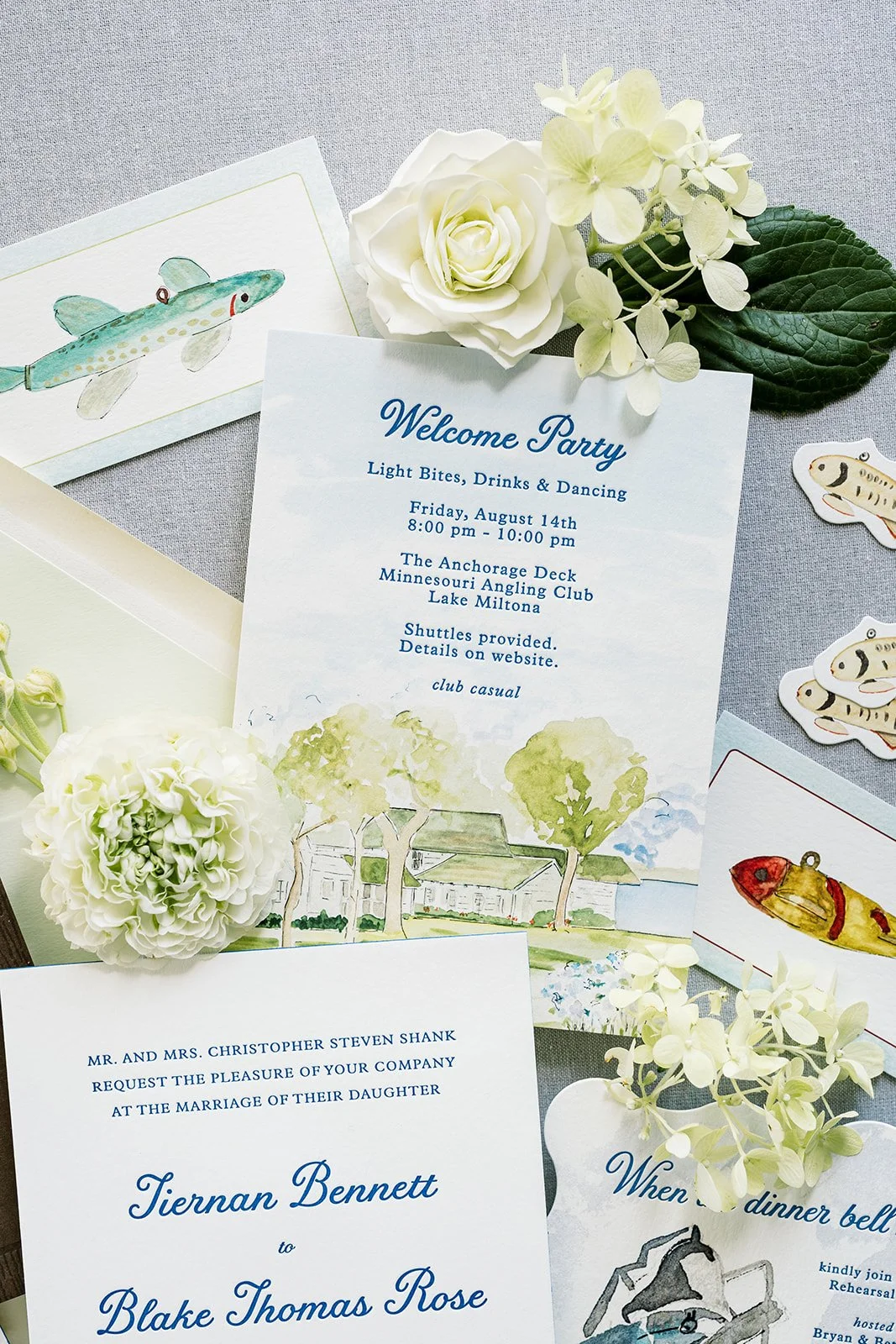 Flat lay of letterpressed and watercolor wedding Invitation and deco items for a fishing-themed welcome party, including flowers and fishing lure cutouts.
