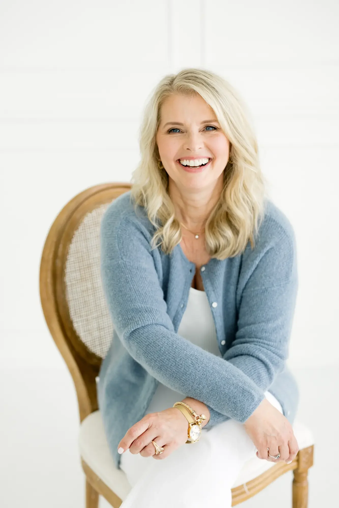 A smiling woman with blonde hair sitting in a wooden chair with a beige cushioned seat, wearing a light blue cardigan with gold jewelry, against a plain white background.