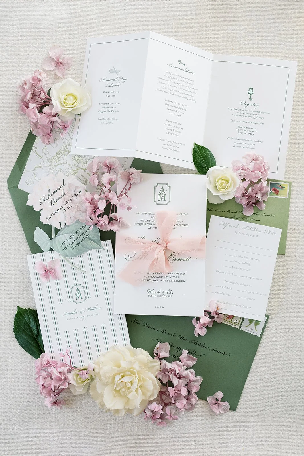 A flat lay of letterpressed wedding and event invitation cards, RSVP cards, and event details on a green envelope, decorated with pink and white flowers and green leaves.