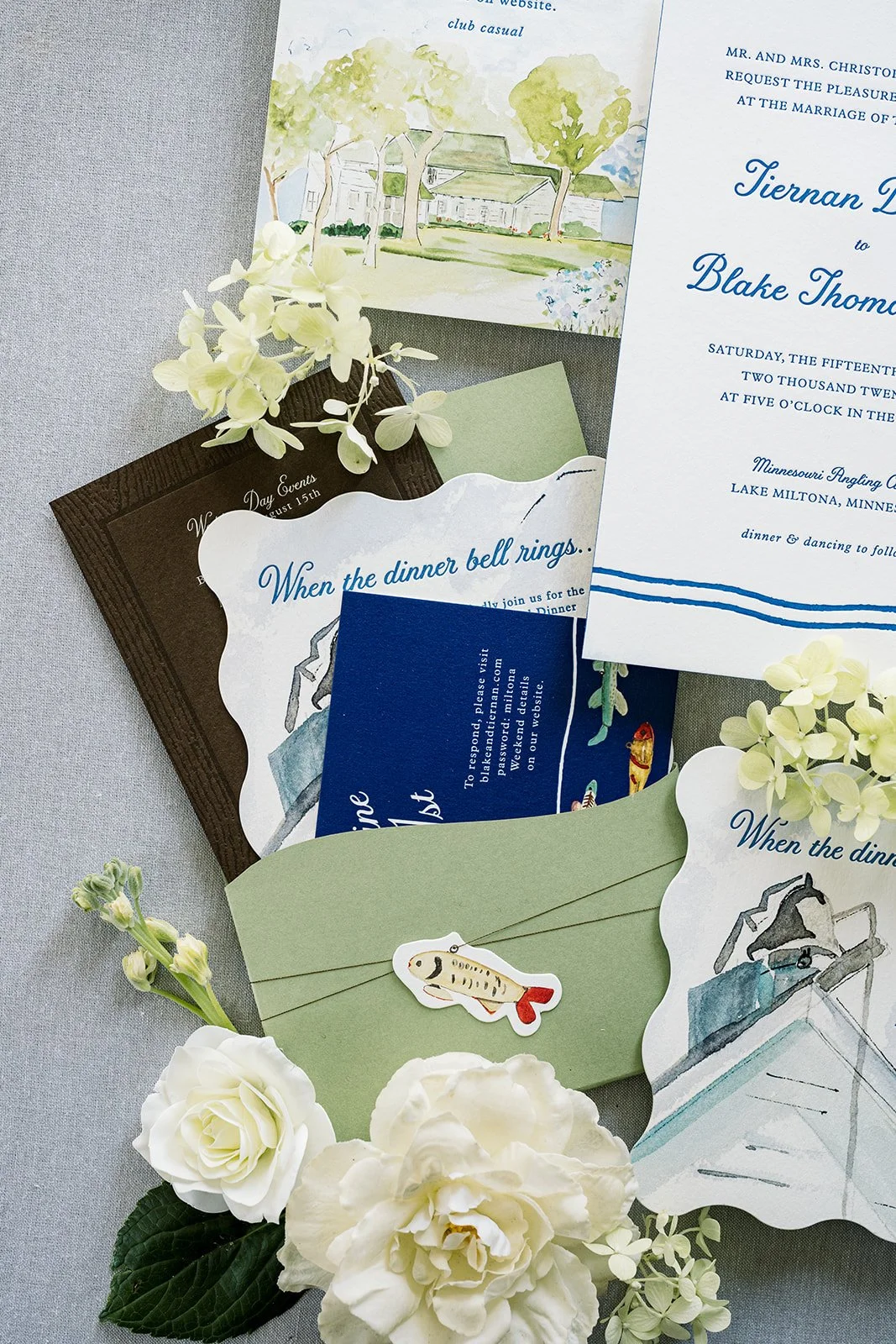 edding invitation suite with floral decorations. The suite includes an illustrated map, an invitation card in white and blue, a brown wedding event card, a response card, and a green envelope with a fish tag. White flowers and greenery surround the stationery.