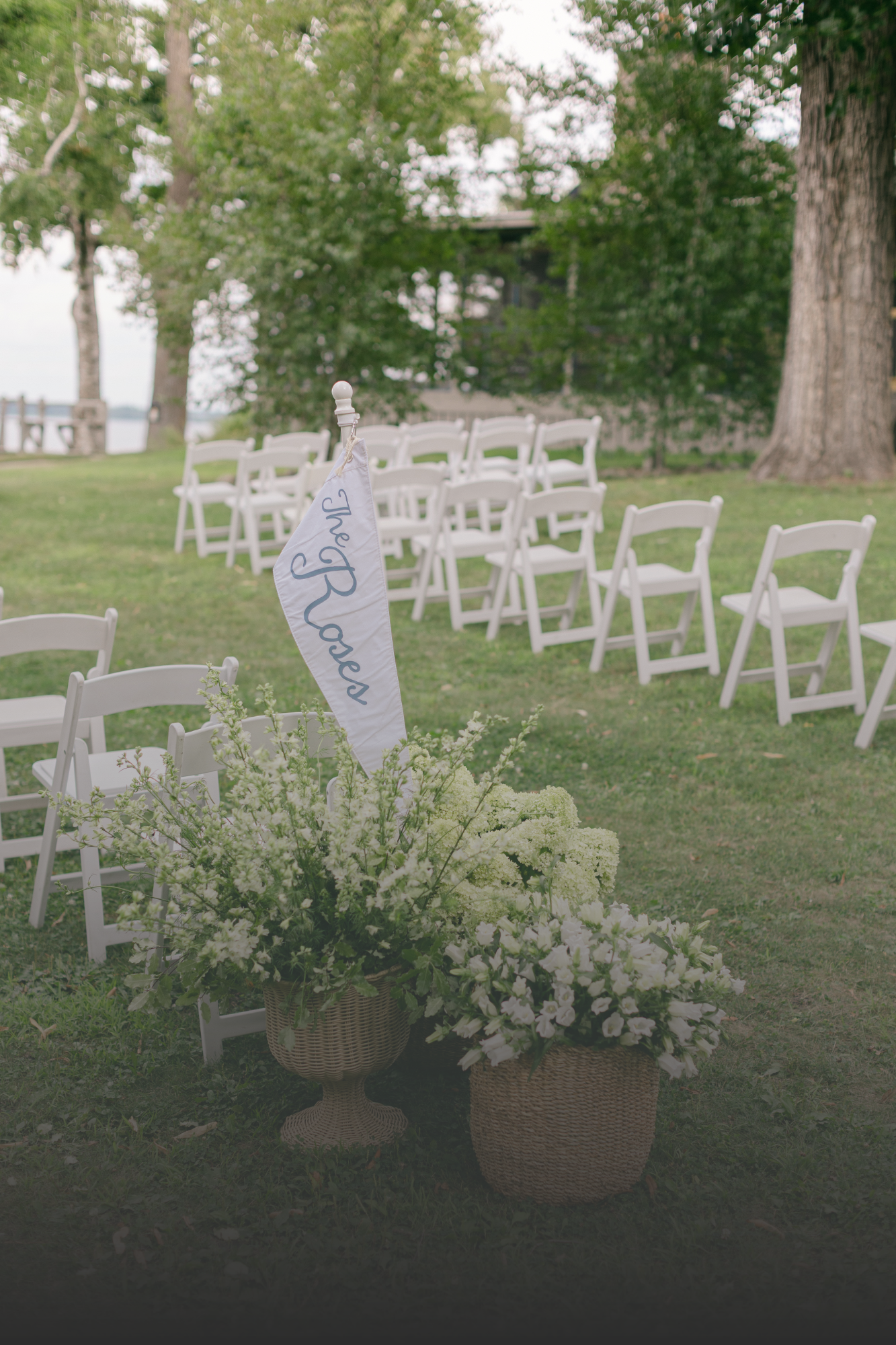 Outdoor wedding setup with white chairs arranged in rows, floral arrangements in baskets, and a white flag that reads 'Yes Please' on a grassy area near trees