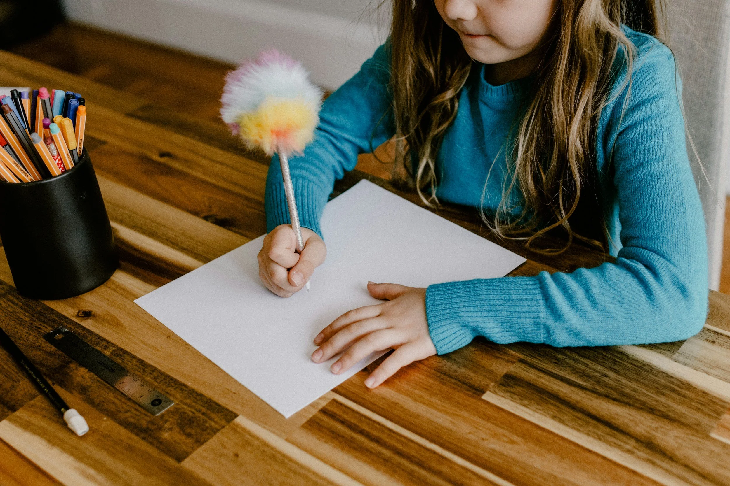 A young girl with long wavy hair wearing a blue sweater sitting at a wooden table, holding a colorful fluffy pen over a blank white sheet of paper. There is a black container filled with various colored pens and a black marker with a white cap on the table.