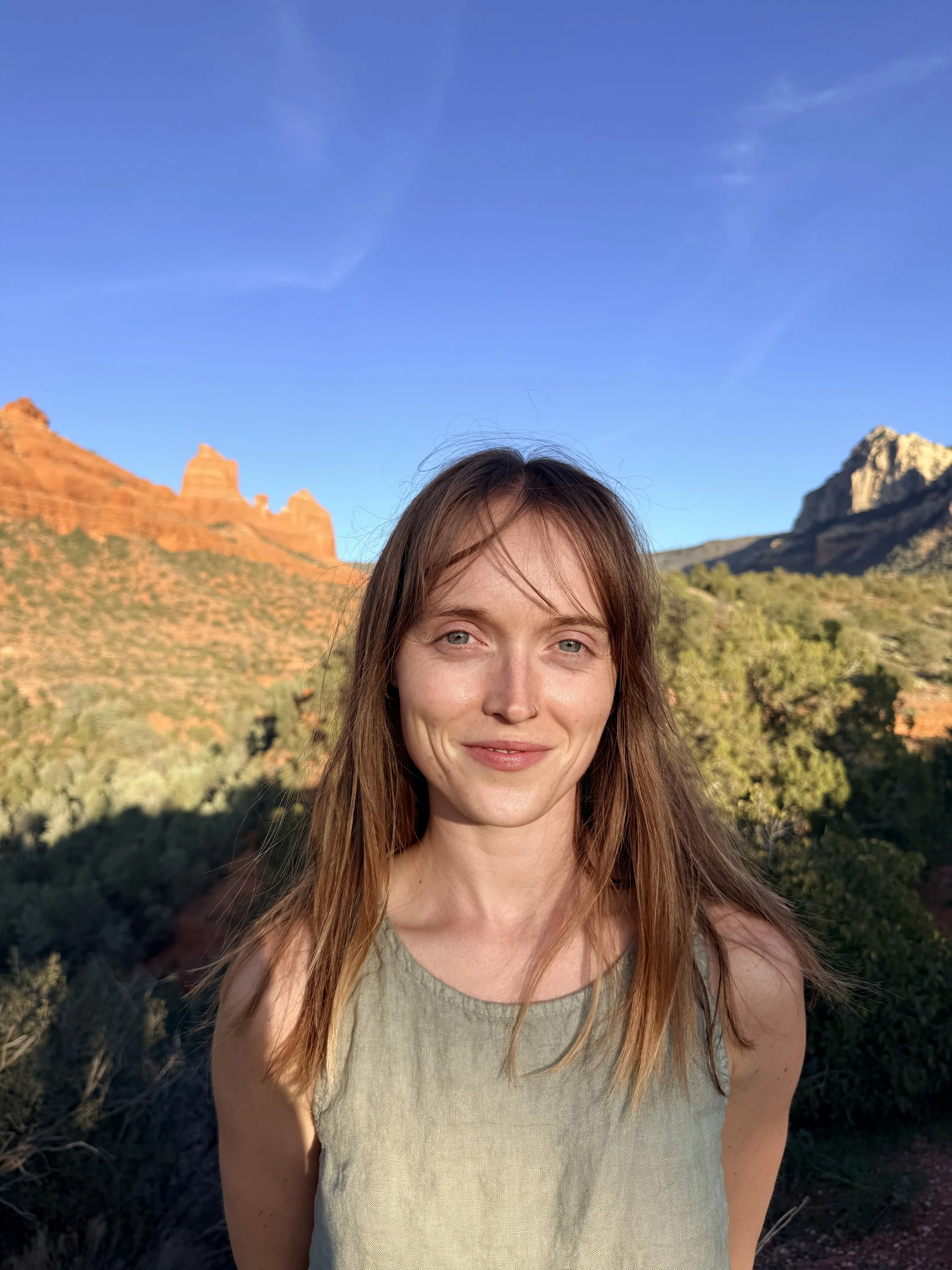 A woman with long reddish-brown hair and blue eyes standing outdoors in front of red rock formations and green vegetation under a clear blue sky.