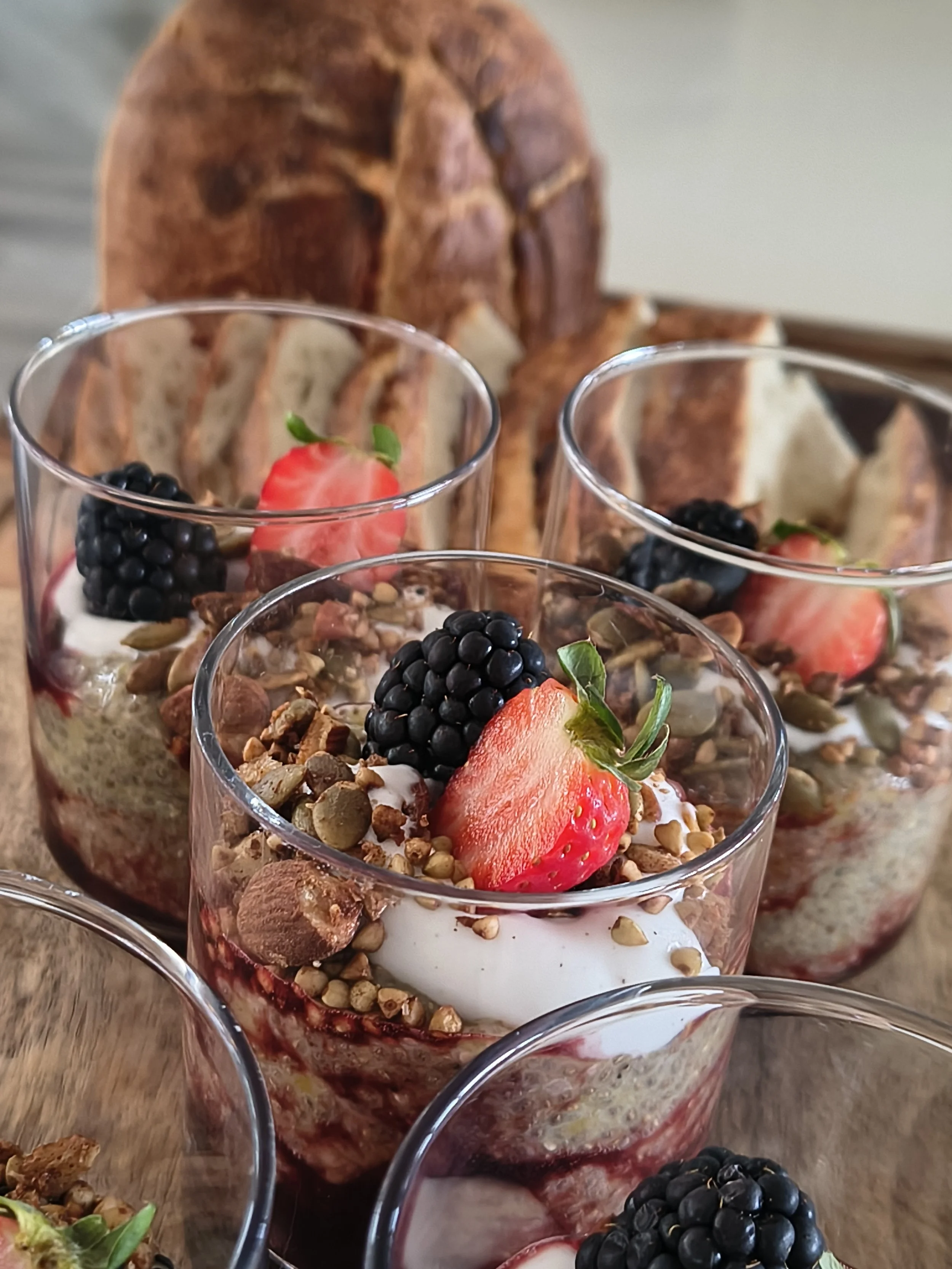 Multiple glasses of layered dessert with strawberries, blackberries, granola, and cream, arranged on a wooden surface with a large, baked loaf of bread in the background.