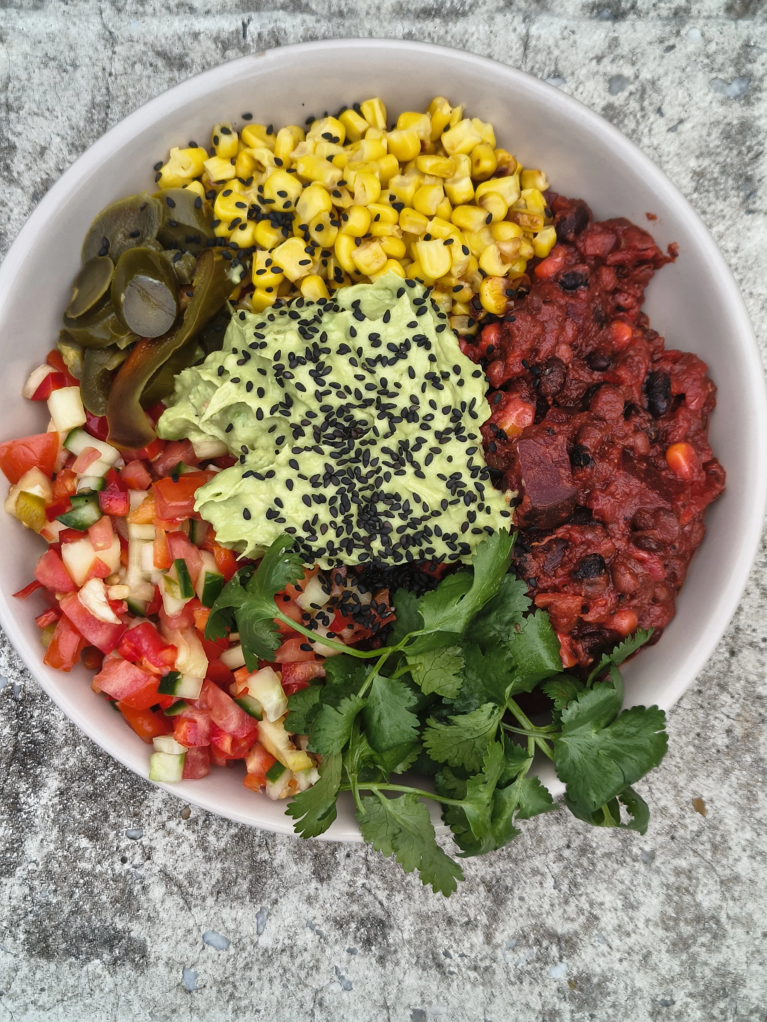 A white bowl with a variety of colorful food items including diced vegetables, guacamole with black sesame seeds, corn, black bean chili, pickles, and cilantro, placed on a textured gray surface.