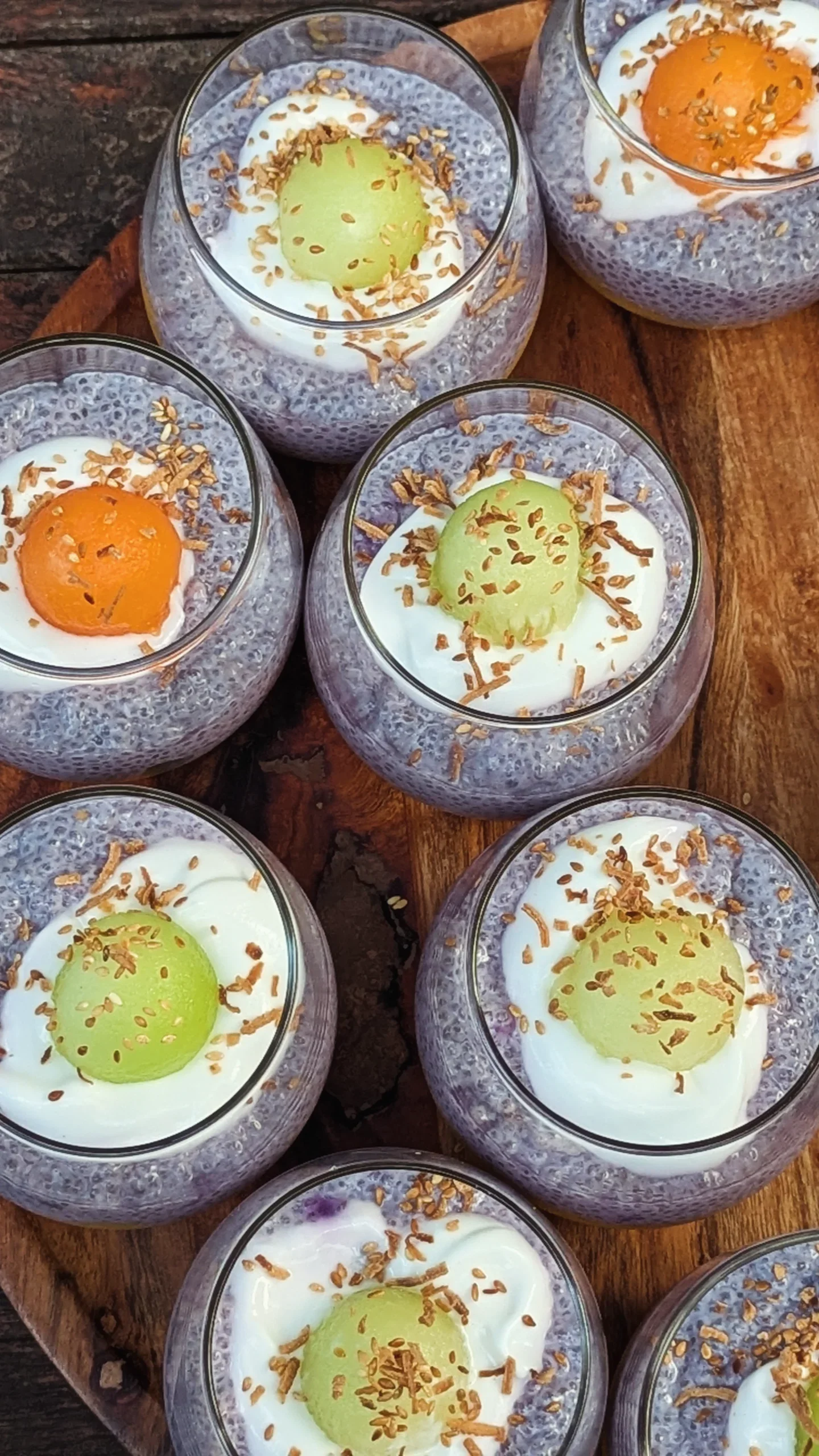 Several glasses filled with chia pudding topped with chopped nuts, garnished with halves of green and orange fruits, served on a wooden surface.