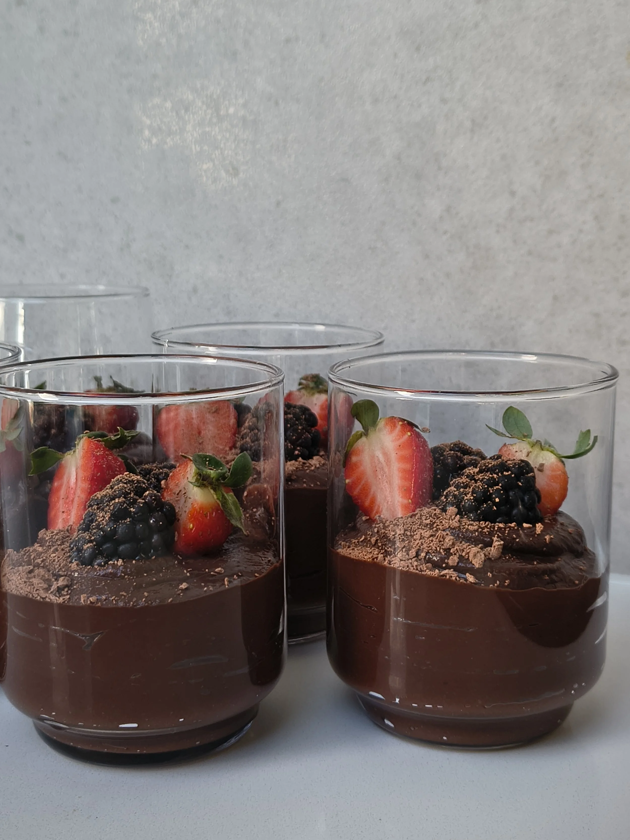Three glasses of chocolate mousse topped with strawberries, blackberries, and green leaves, on a white surface.