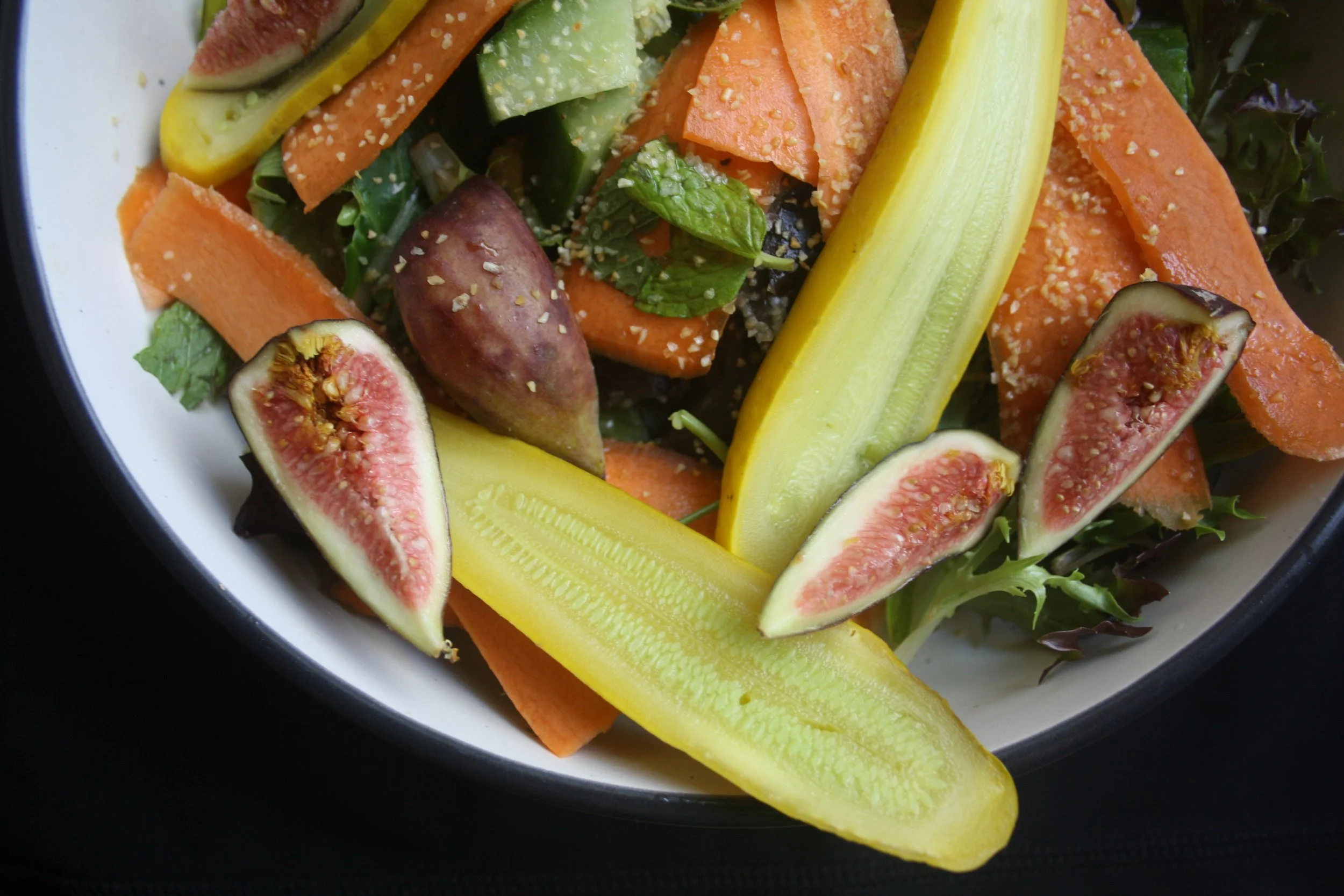 Close-up view of fresh vegetables including sliced figs, yellow and orange bell peppers, carrots, cucumbers, purple potatoes, and leafy greens in a white bowl.
