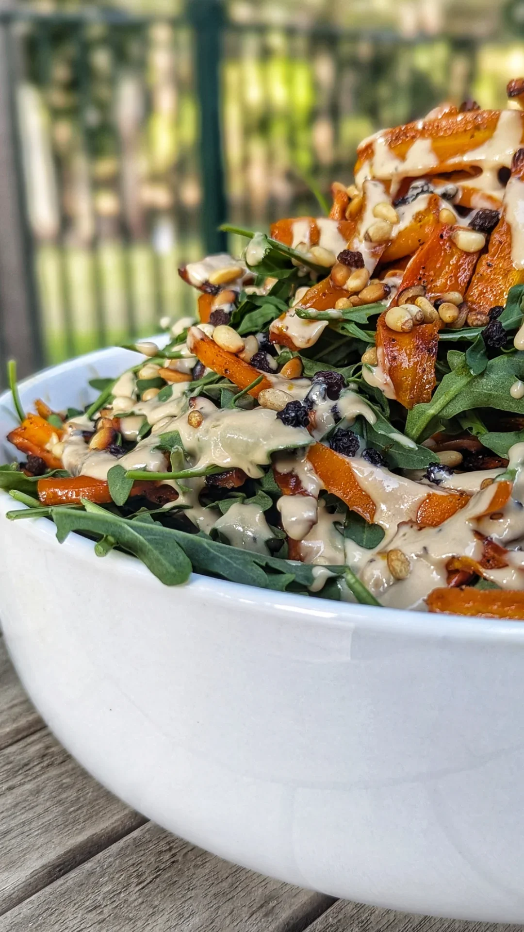 A white bowl filled with a mixed salad featuring arugula, roasted sweet potatoes, pine nuts, dried cranberries, and drizzled with a creamy dressing.