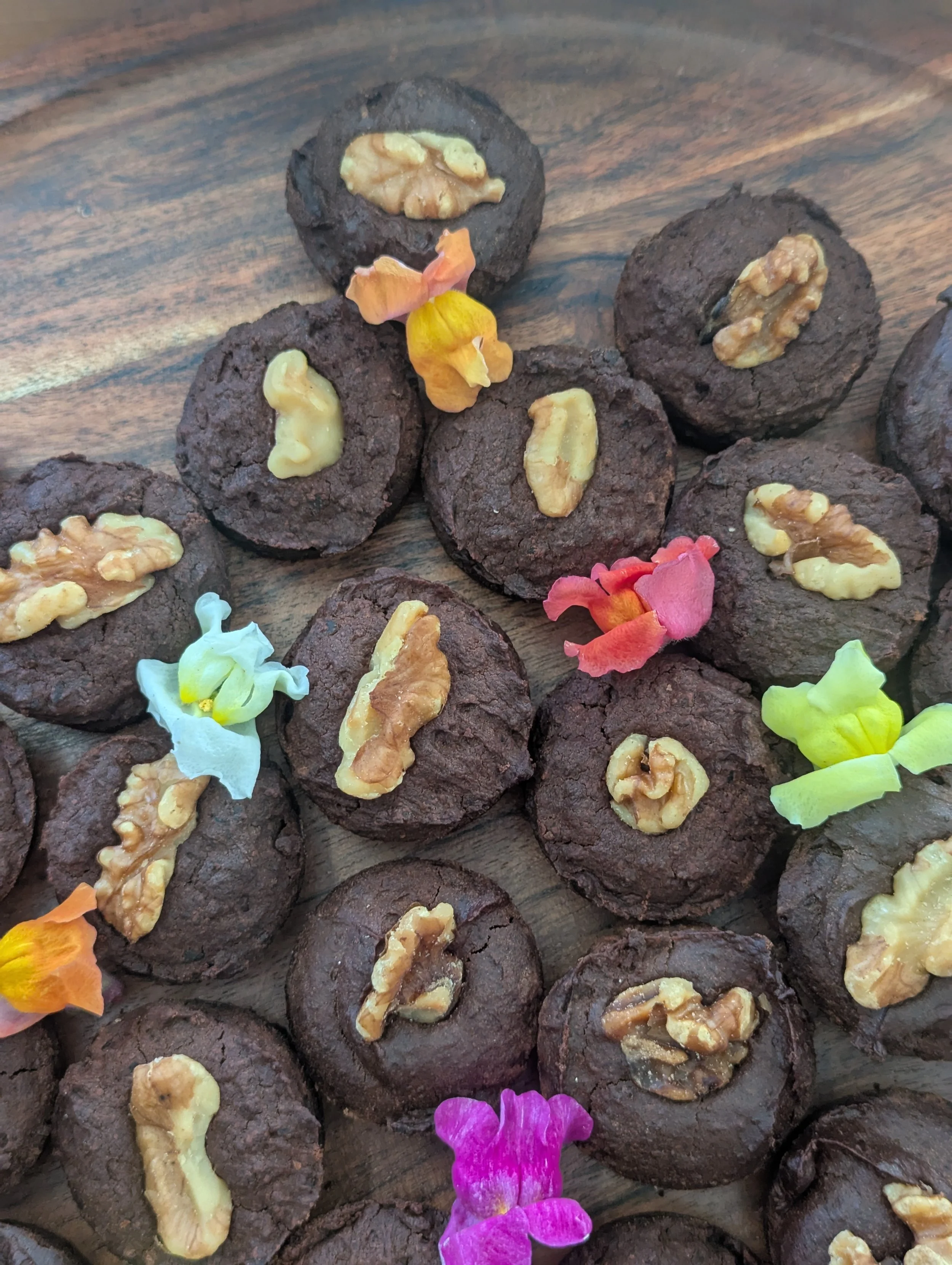 Several round, dark chocolate cookies topped with walnuts, displayed on a wooden surface alongside colorful flower petals.