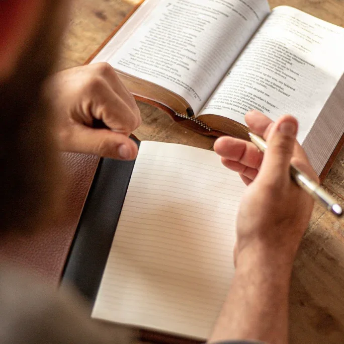Person taking notes in a notebook with an open book nearby on a wooden surface.