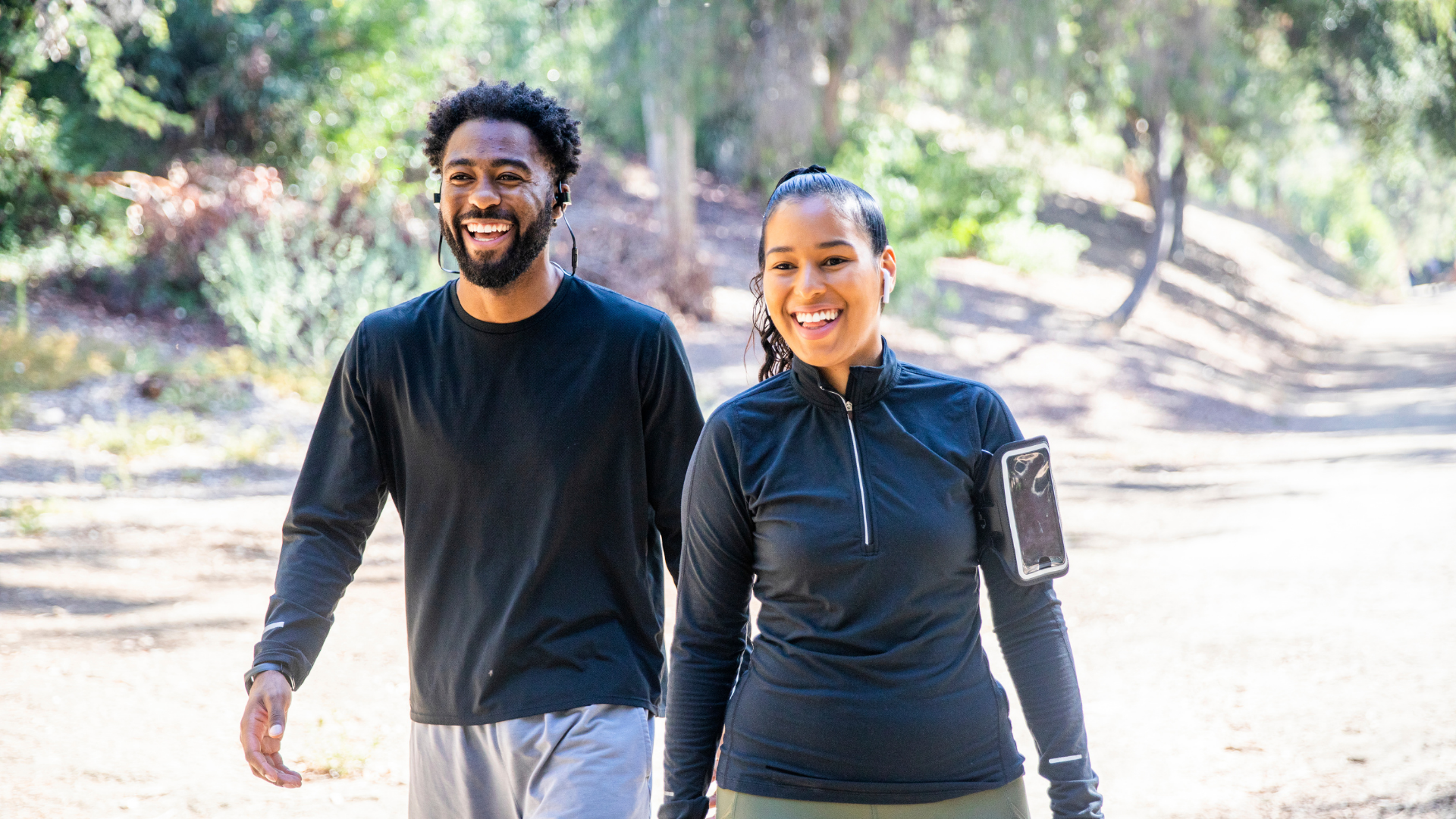 A man and woman are walking outdoors on a trail in a forested area, both smiling and wearing athletic clothing and earphones.