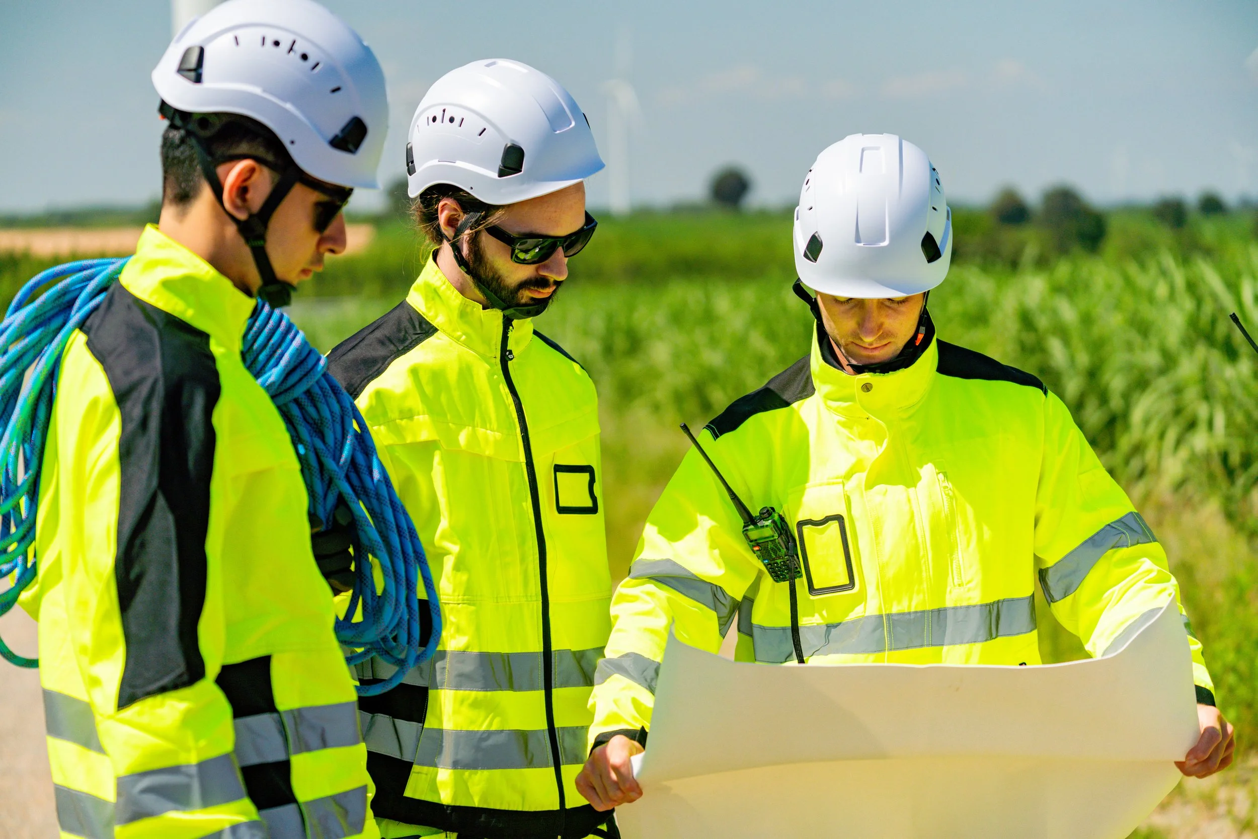 A team of environmental consultants examining plans while dressed in safety gear under a clear sky in a rural area.