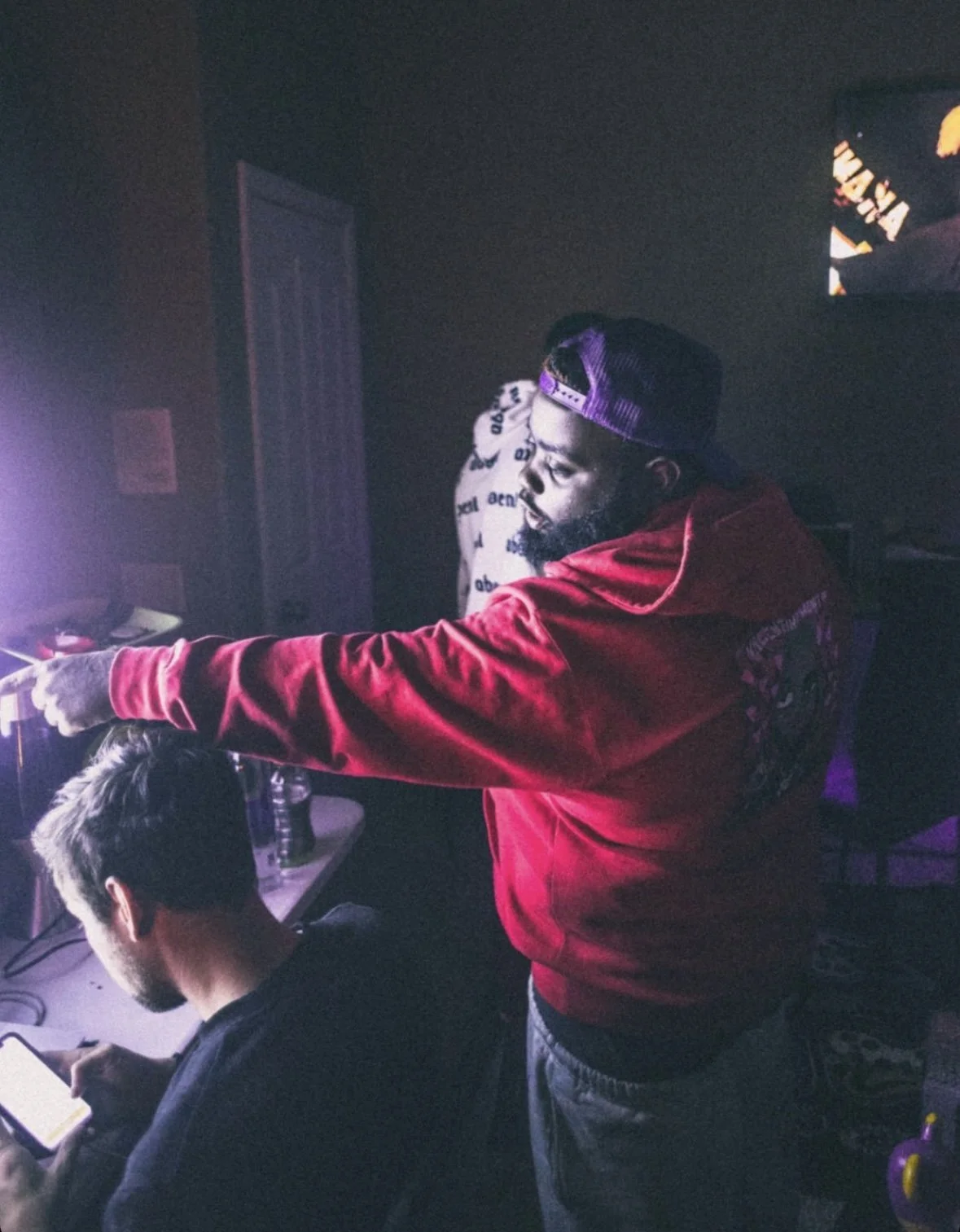 A man with a beard and a purple cap points at a computer screen in a dimly lit room with another man using a phone nearby, and a television on the wall.