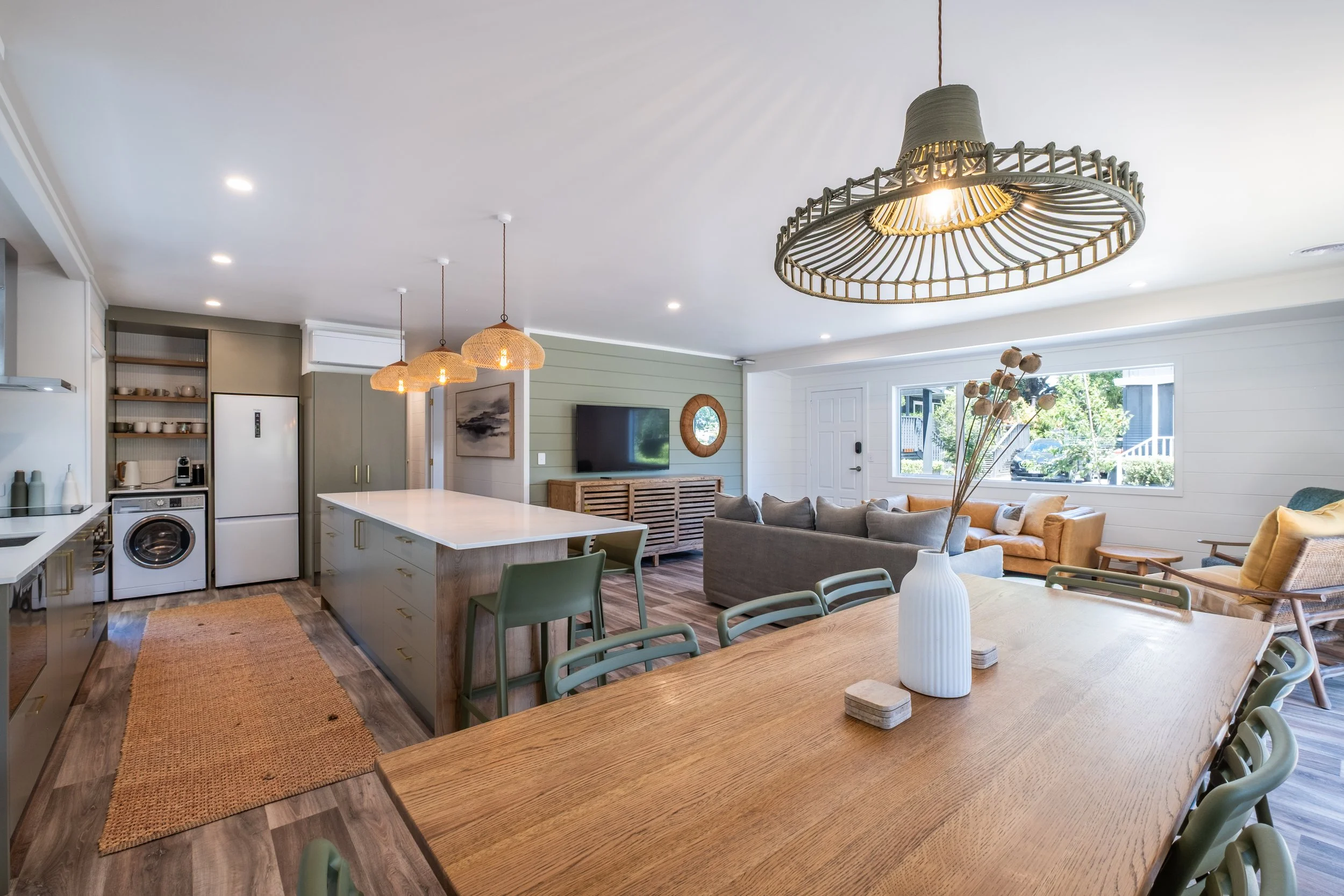 Open-concept living space with a kitchen, dining area, and living room, featuring modern decor, light-colored walls, wood accents, and large windows letting in natural light.
