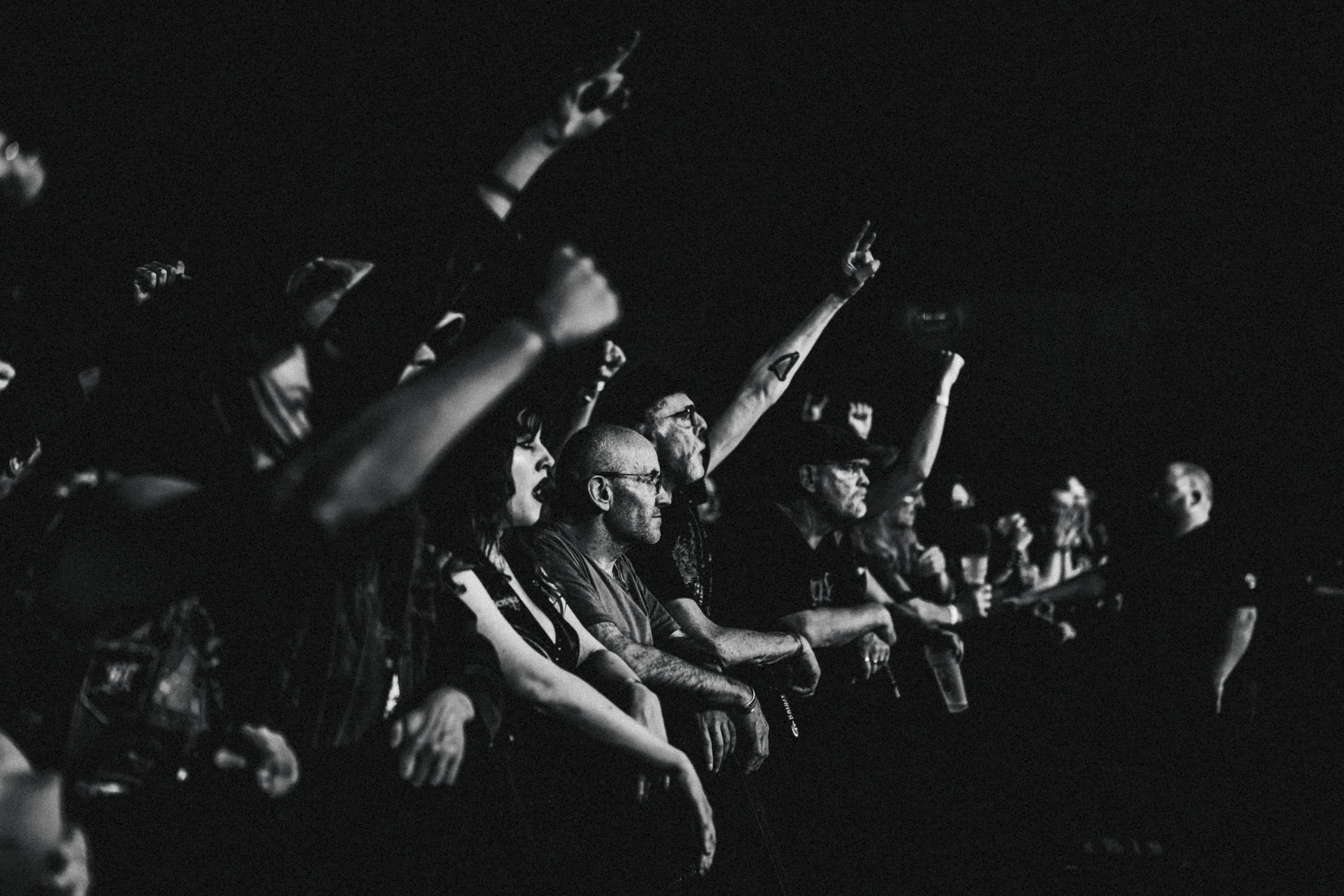 Groupe de personnes assises dans une salle sombre lors d'un événement ou d'un concert, certaines levant les mains, en noir et blanc.