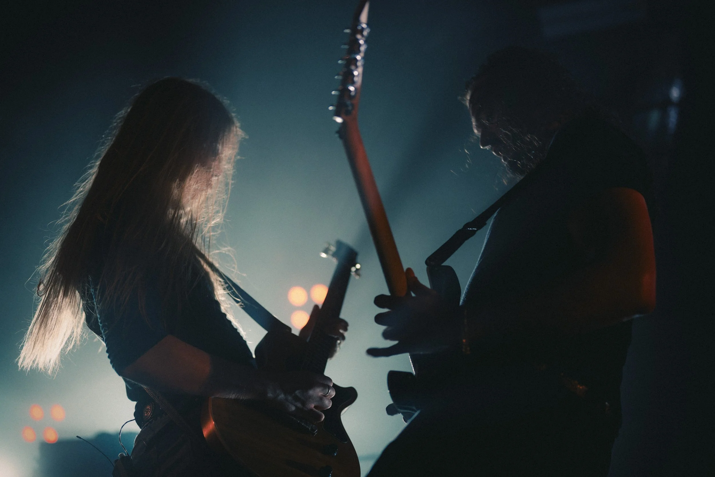 Deux personnes jouent de la guitare électrique dans une ambiance sombre et éclairée par des lumières d'ambiance.