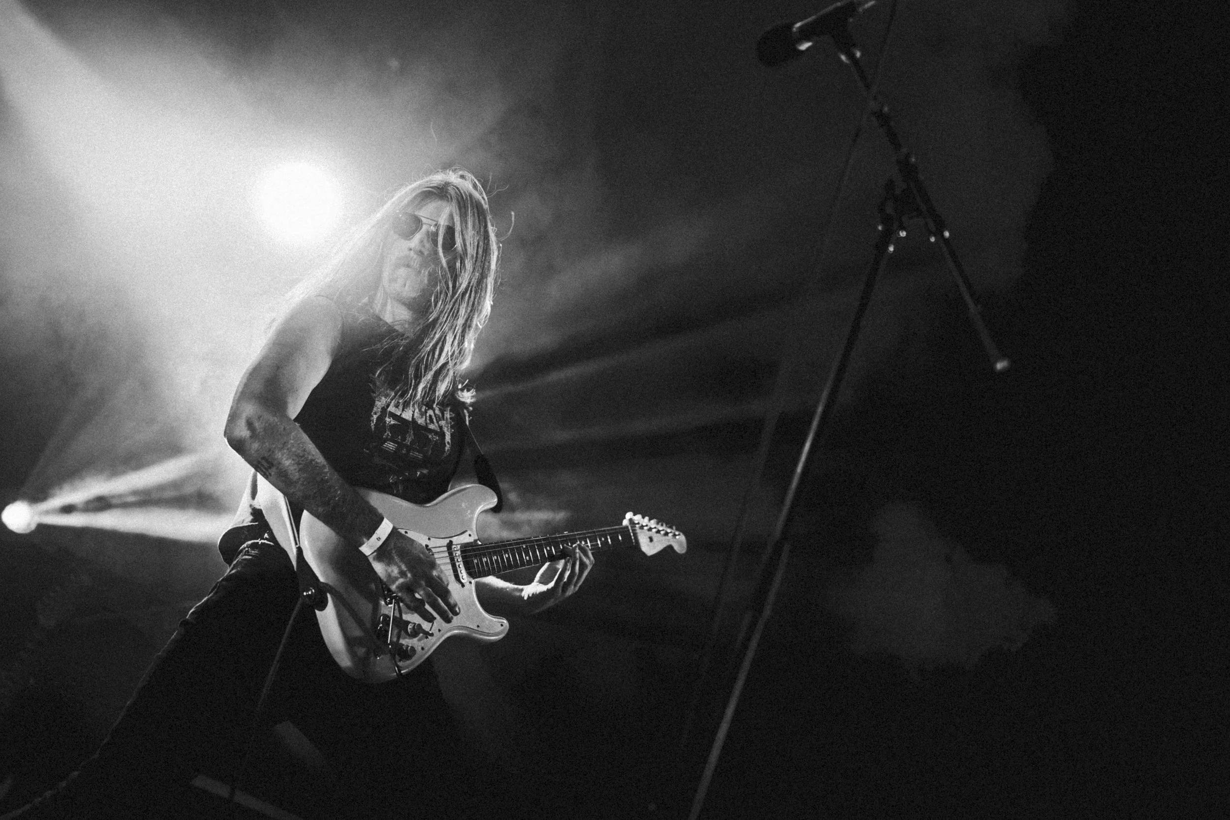 Un musicien joue de la guitare électrique sur scène lors d'un concert, avec des lumières de scène en arrière-plan en noir et blanc.