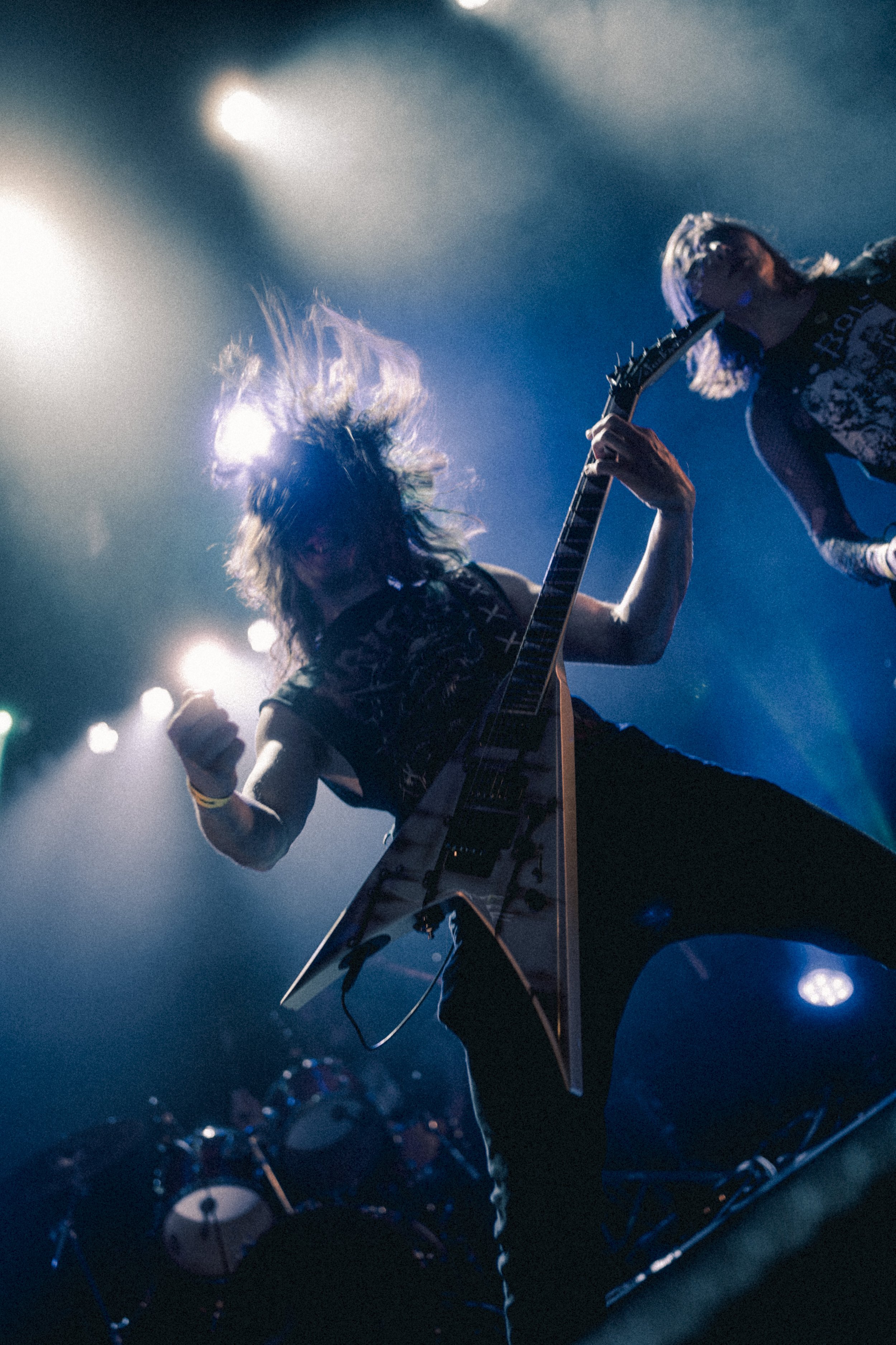 Musicien jouant de la guitare électrique en concert, avec cheveux en mouvement et lumière colorée en arrière-plan.