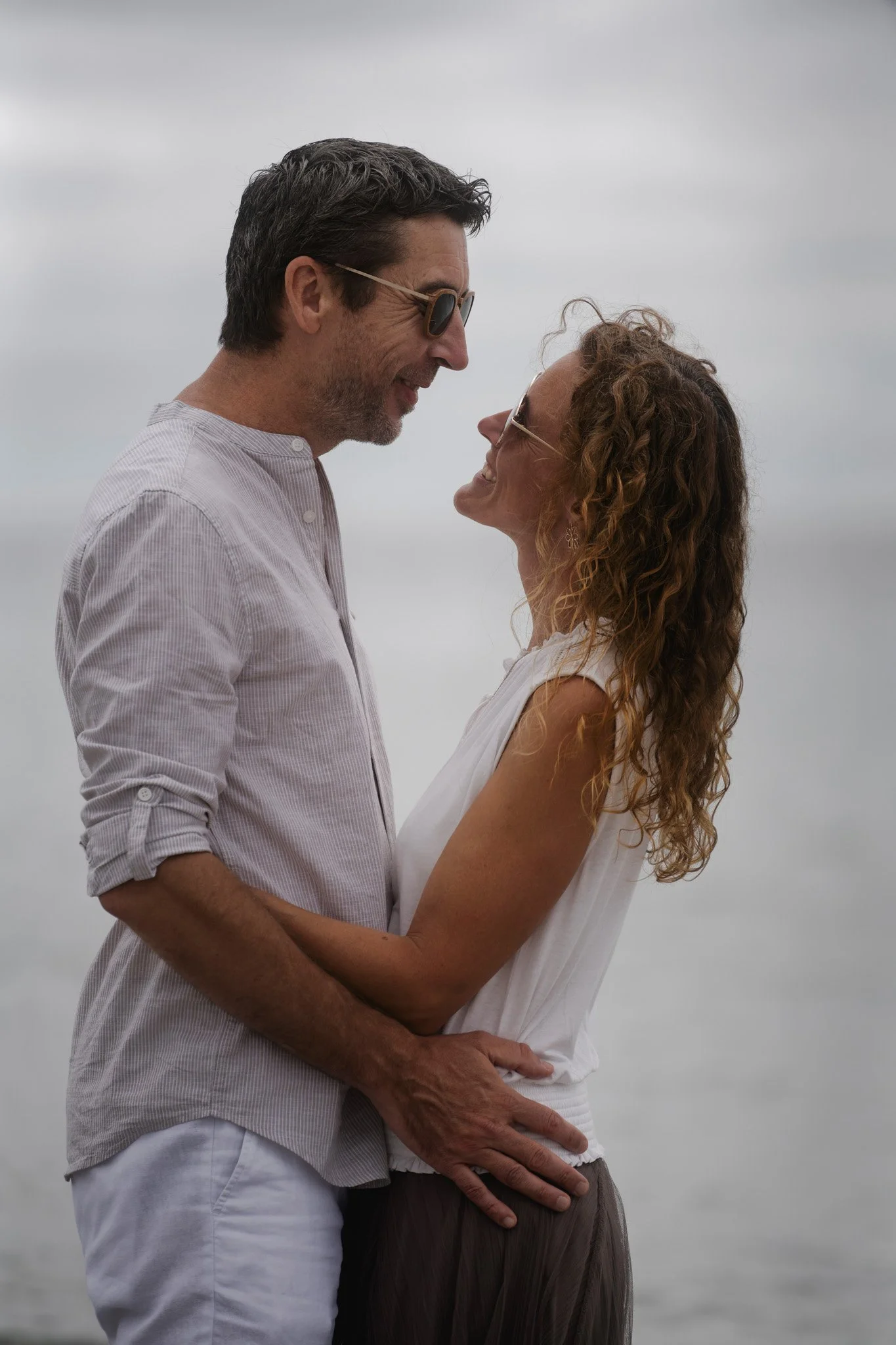Un couple qui se tient face à face près de la plage, souriant et regardant l'un l'autre, par une journée nuageuse.