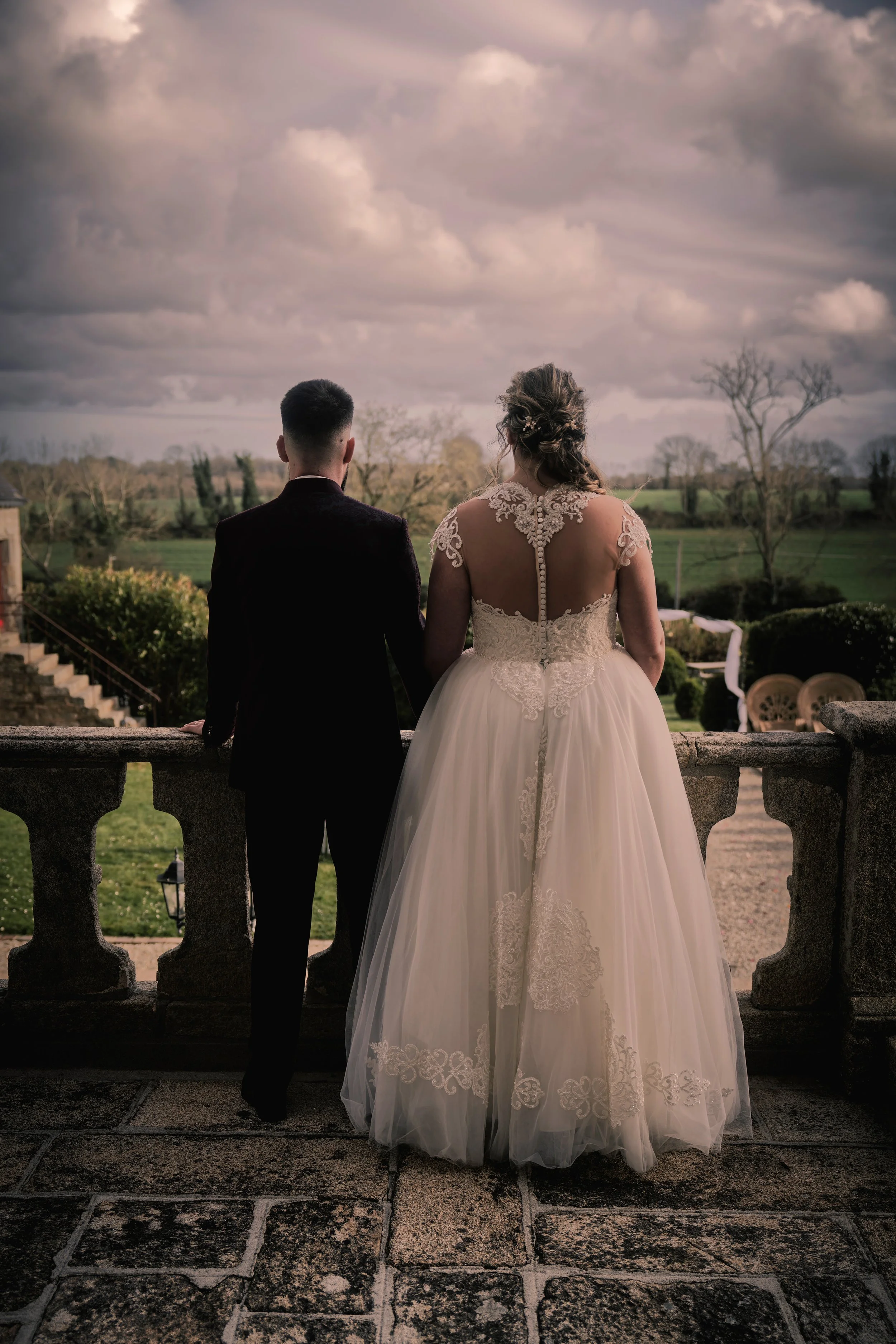 Un couple de mariés vu de dos, debout sur une terrasse en pierre, regardant un paysage rural avec des champs verts et quelques arbres. La mariée porte une robe de mariée blanche avec des détails en dentelle, et le marié porte un costume noir.
