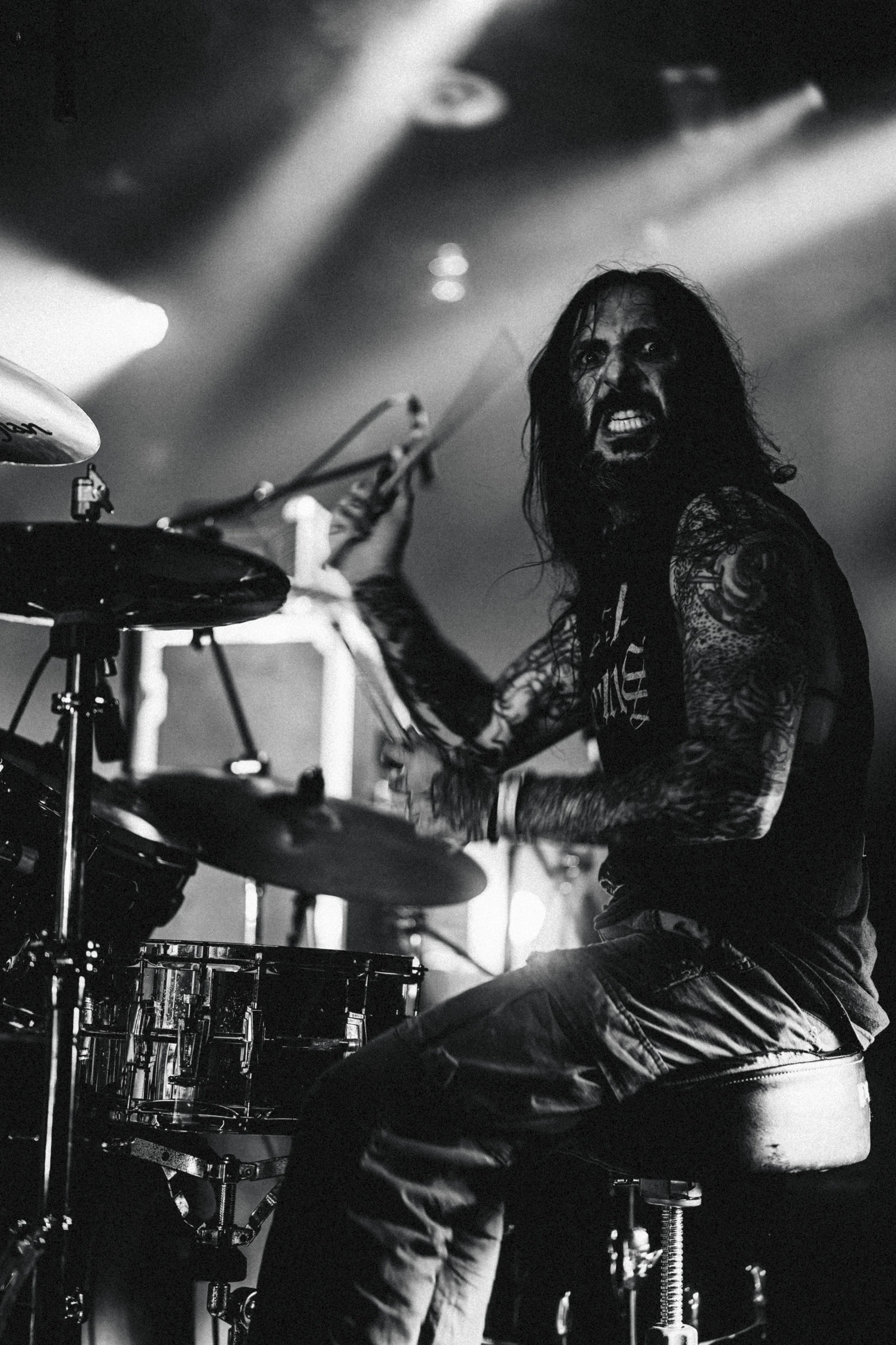 Musicien jouant de la batterie lors d'un concert, en noir et blanc, avec un look intense et musclé, cheveux longs, tatouages visibles.