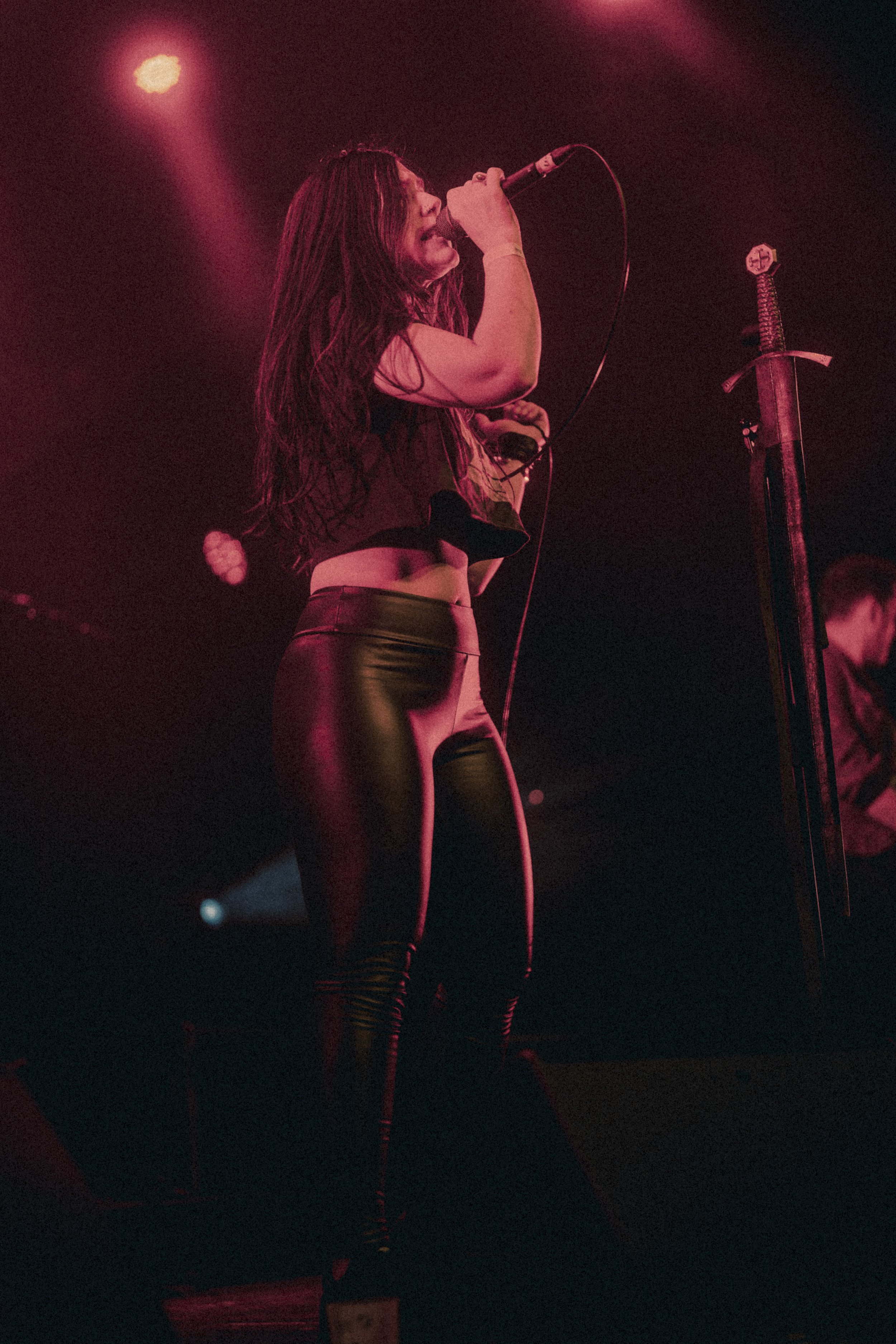 Une chanteuse avec de longs cheveux noirs, portant un haut noir et un pantalon en cuir noir, chante dans un concert sous un éclairage rouge.