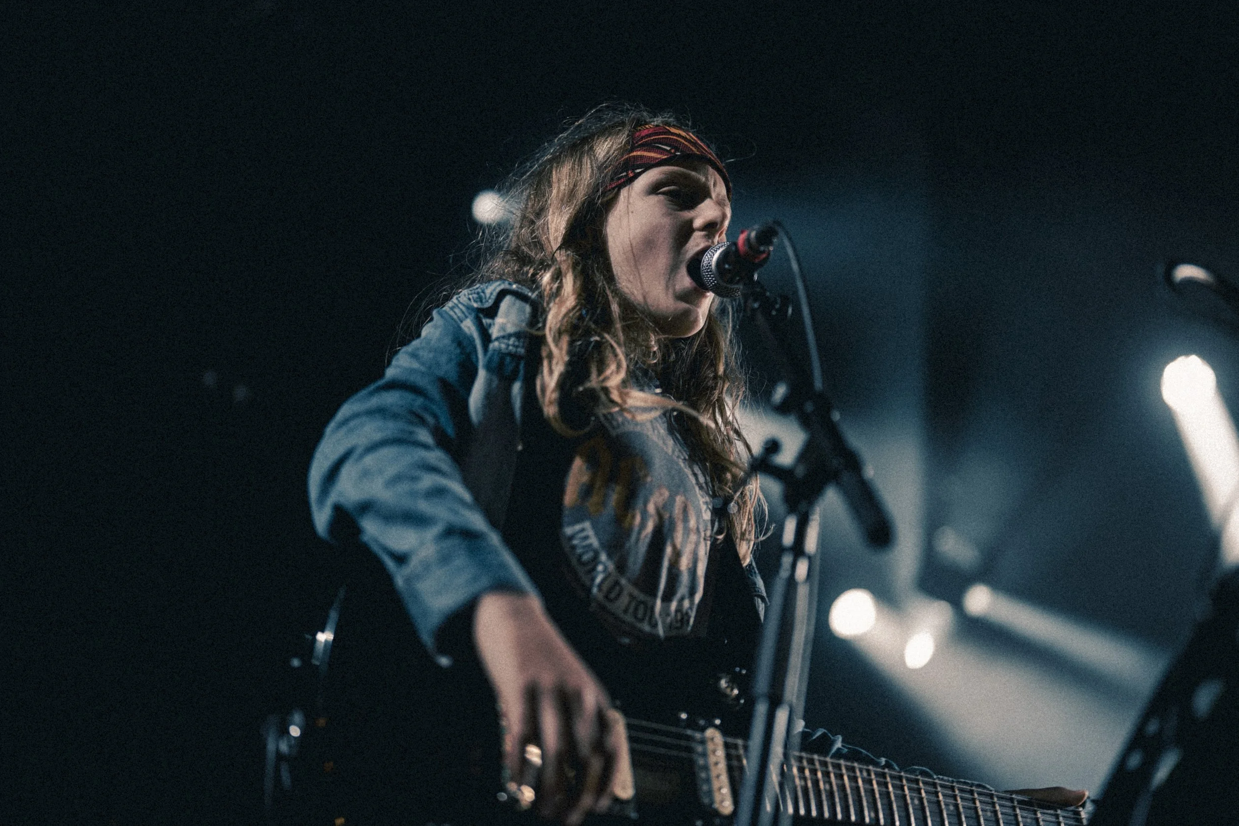 Une femme chante en jouant de la guitare électrique sur scène, portant un bandana rouge et une veste en jean, avec des lumières de scène en arrière-plan.