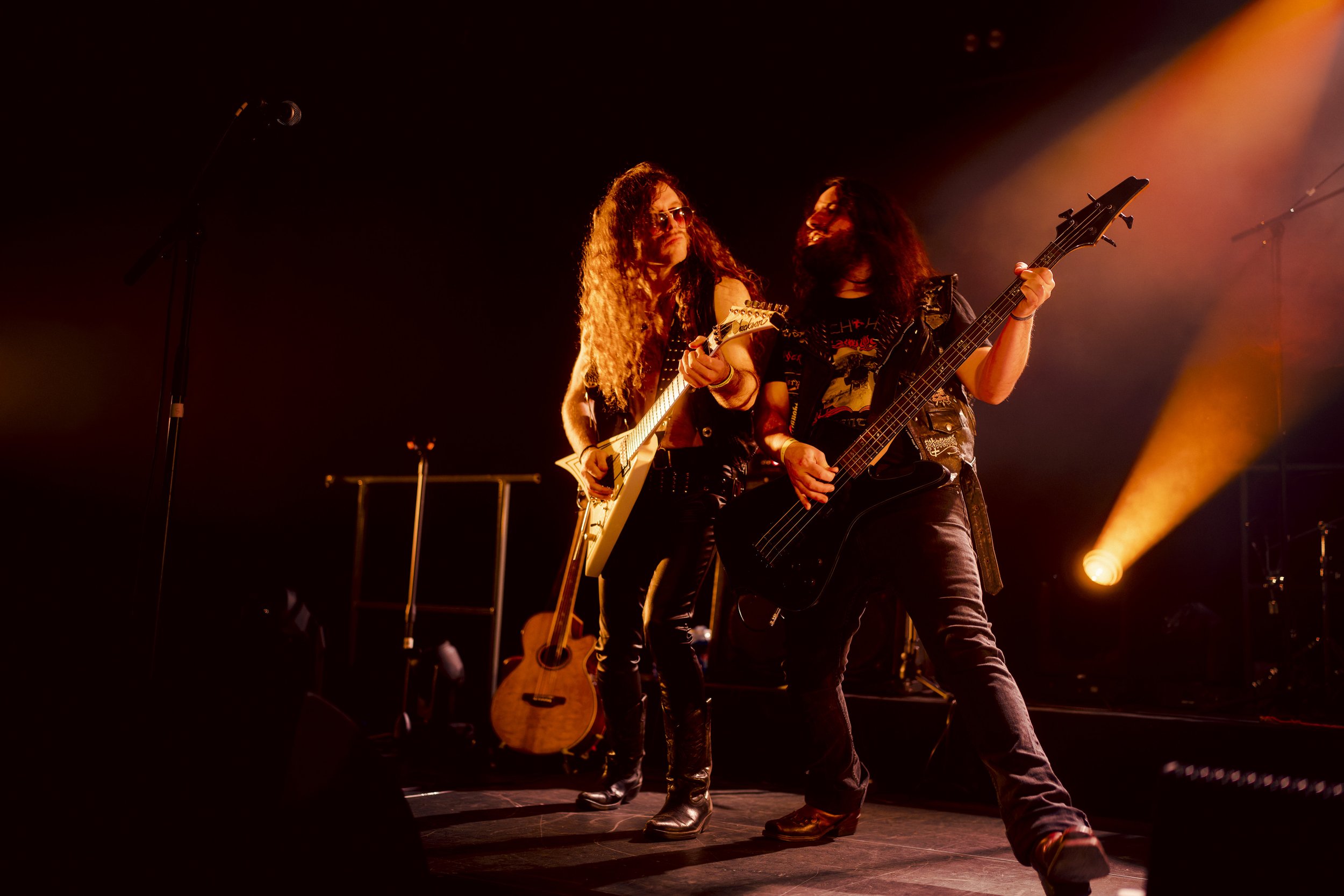 Deux musiciens jouent de la guitare électrique sur scène, sous des lumières orange, cheveux longs, vêtements de style rock.