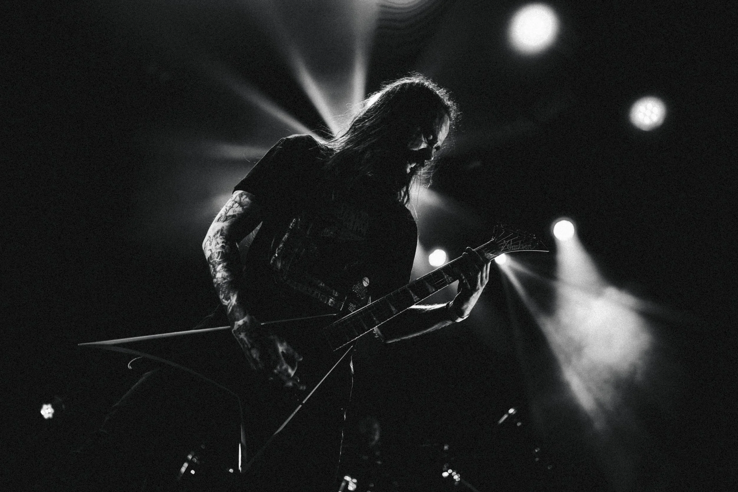 Musicien de rock en noir et blanc jouant de la guitare électrique sur scène avec un rétroéclairage intense derrière lui.