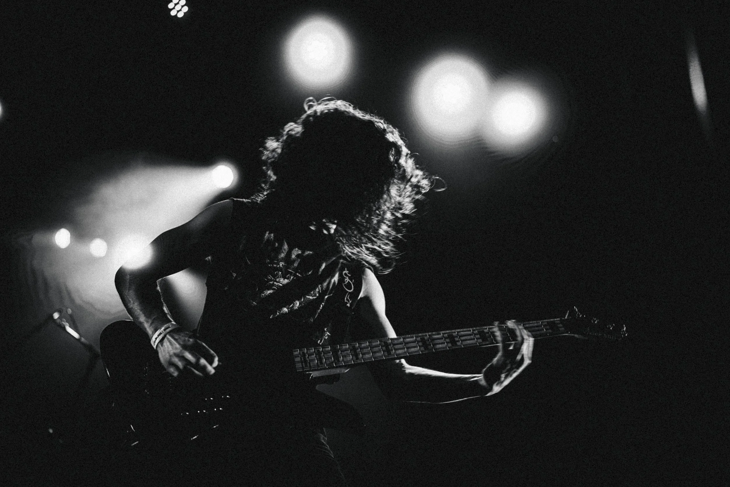 Musicien jouant de la guitare électrique lors d'un concert, silhouettes en noir et blanc, scène sombre avec lumières floues en arrière-plan.