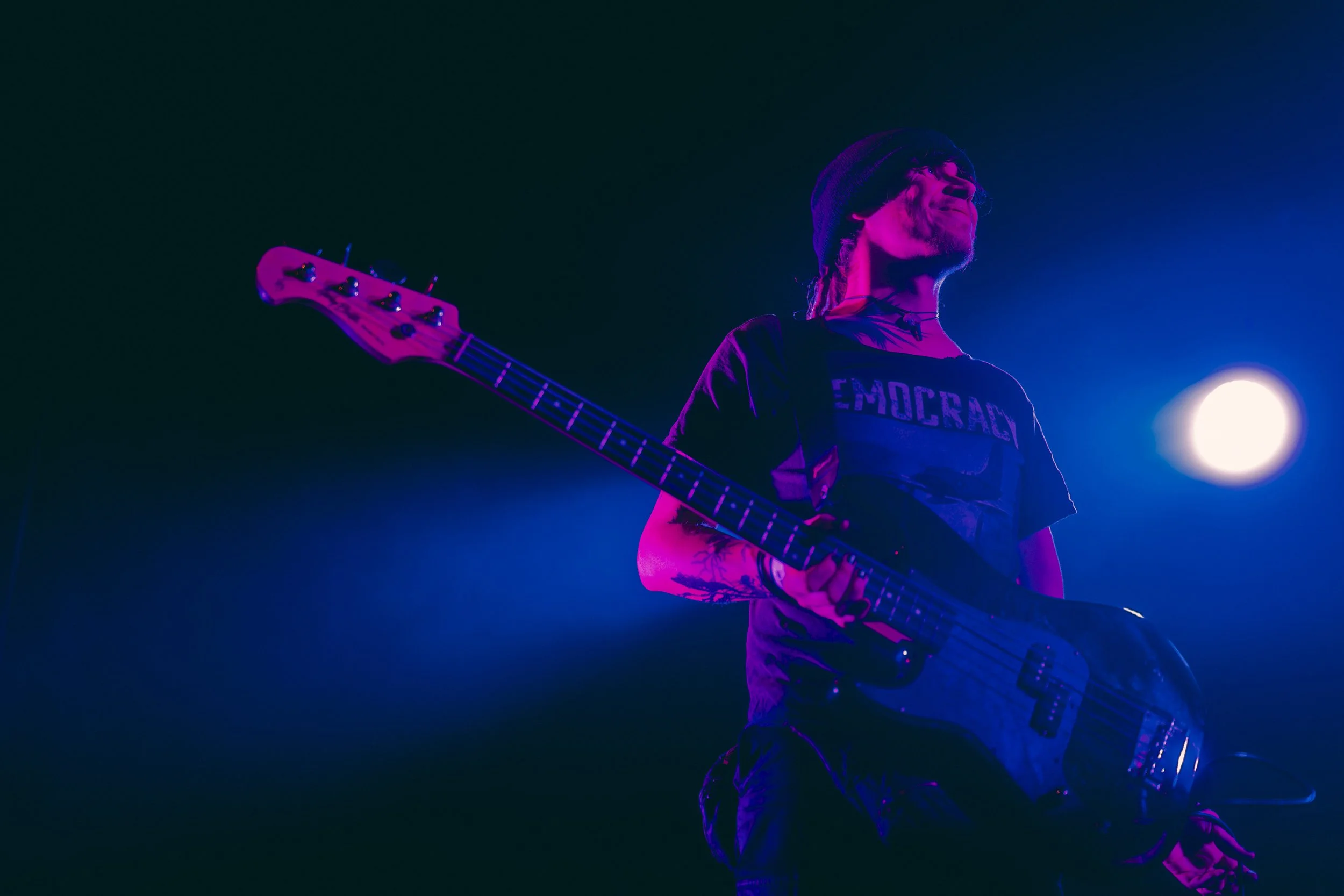 Musicien jouant de la guitare électrique sur scène, éclairage violet et bleu, fond sombre.