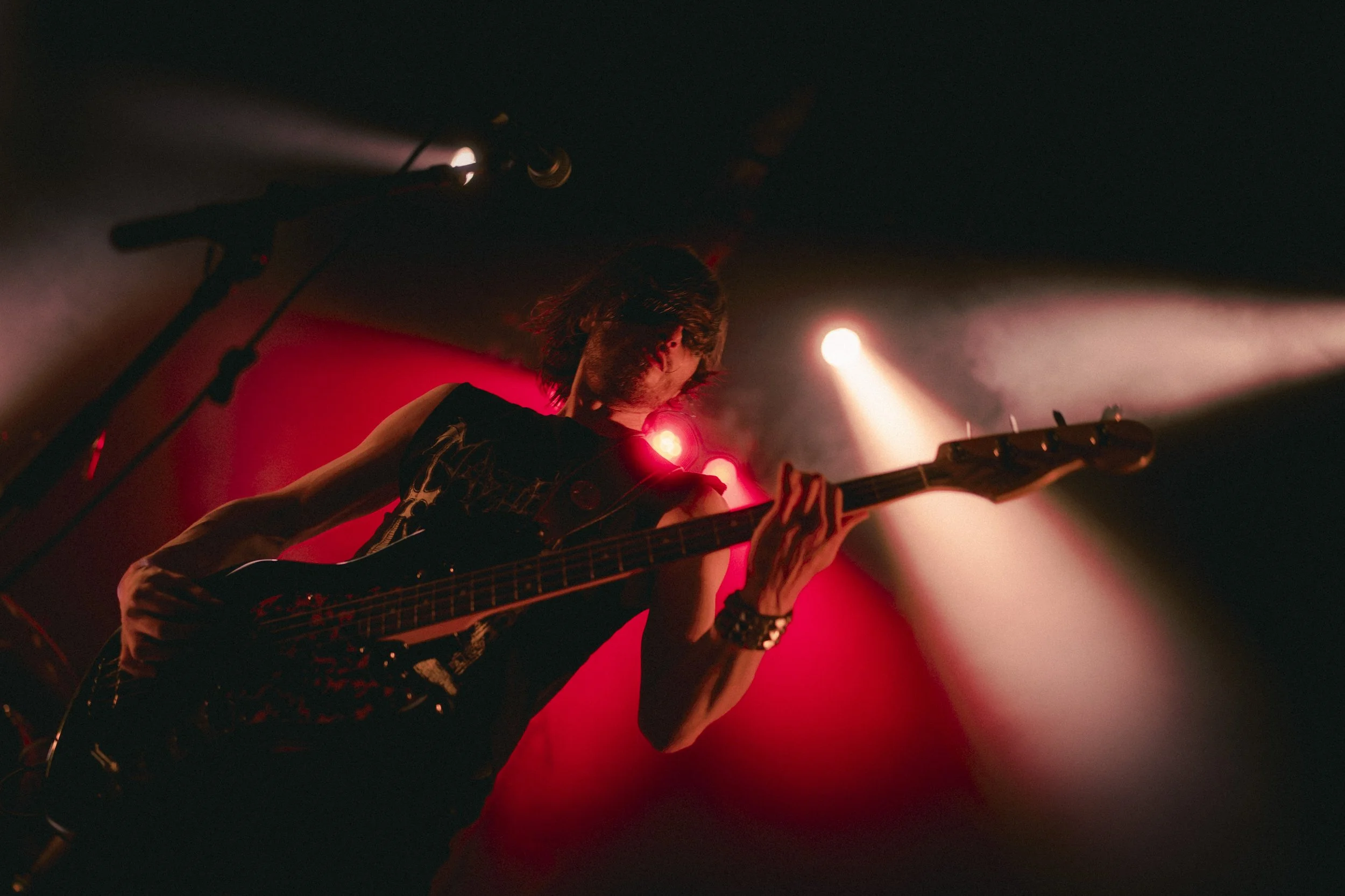 Un musicien joue de la basse électrique lors d'un concert, éclairé par des lumières rouges et blanches dans une atmosphère sombre.