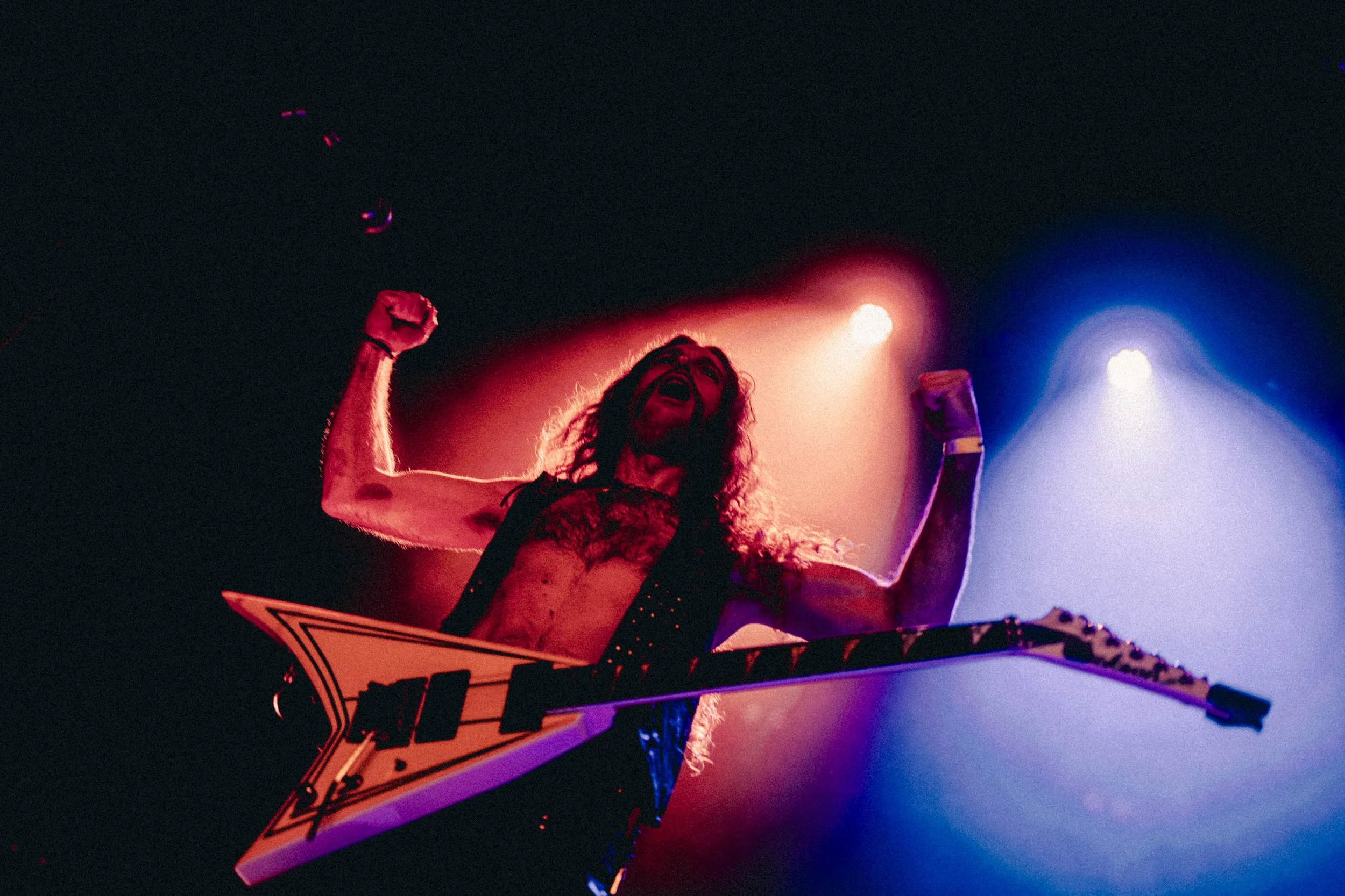Un homme musclé avec des cheveux longs jouant une guitare électrique en forme de losange sur scène, avec des lumières colorées rouge, violet et bleu en arrière-plan, exprimant une émotion intense.