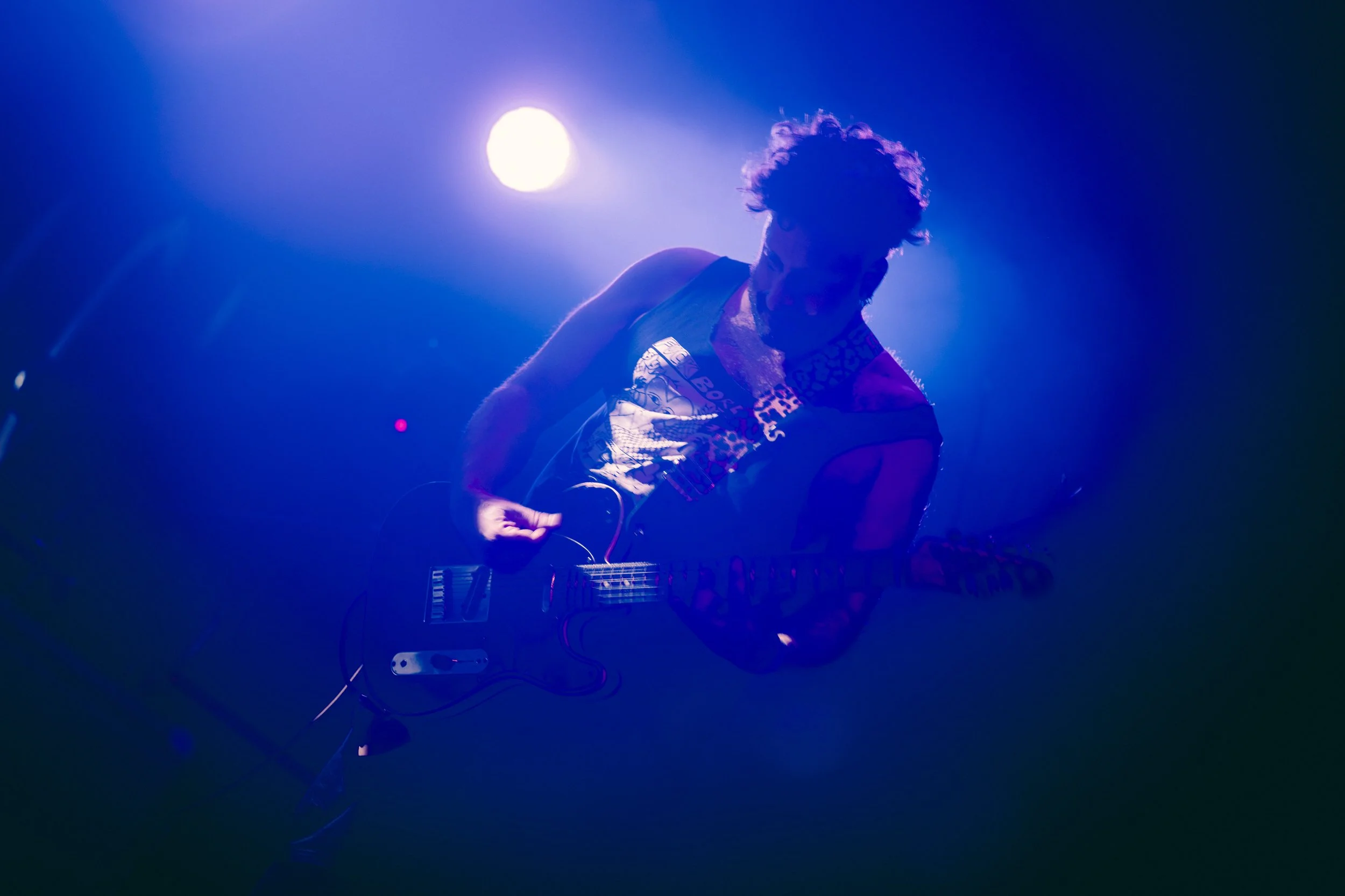 Musicien jouant de la guitare en scène sous un éclairage violet et blanc.