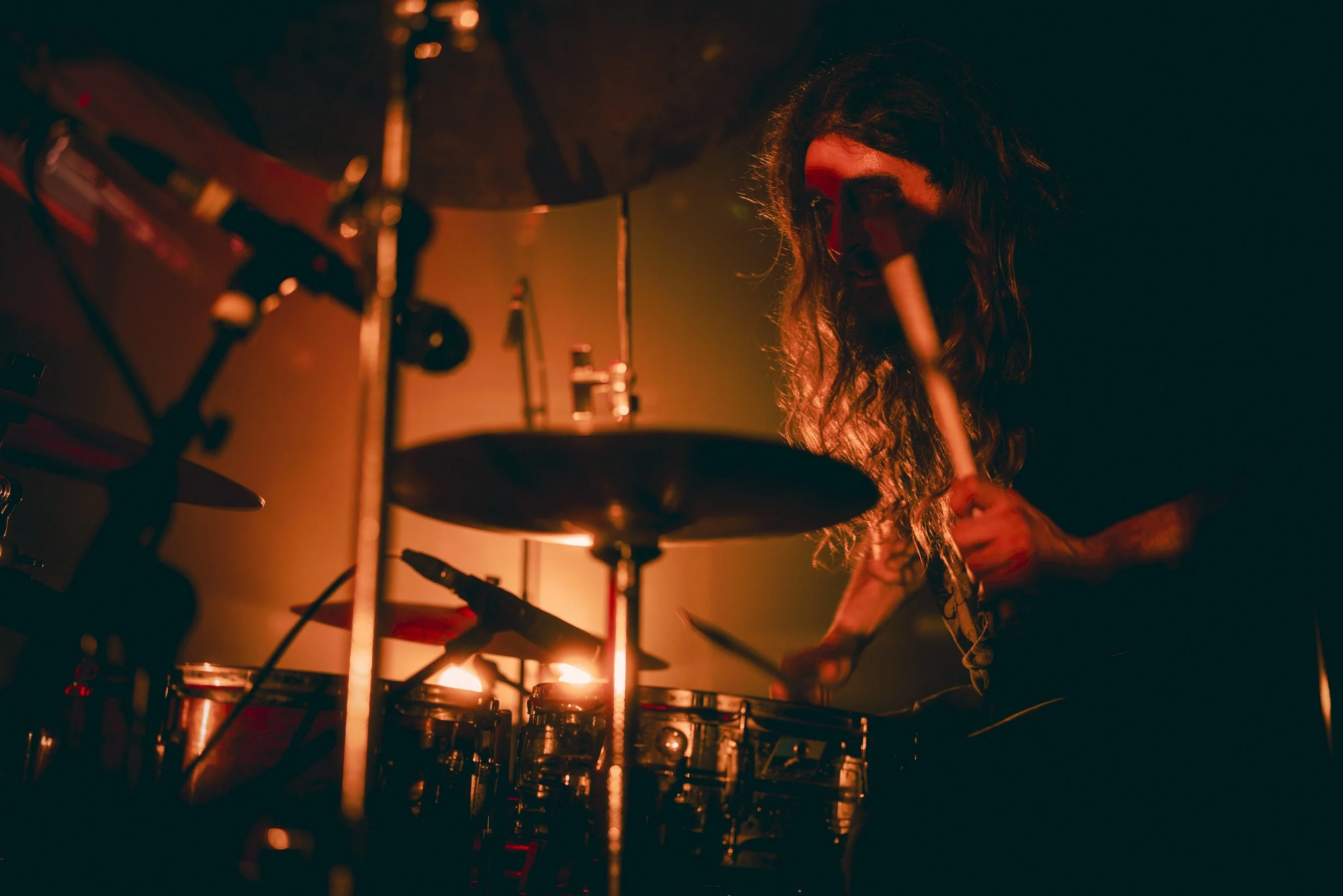 Un musicien avec de longs cheveux joue de la batterie dans une ambiance sombre et rougeâtre.