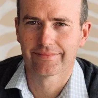 Close-up of a man with short dark hair, light skin, wearing a checkered shirt and blazer, smiling at the camera.