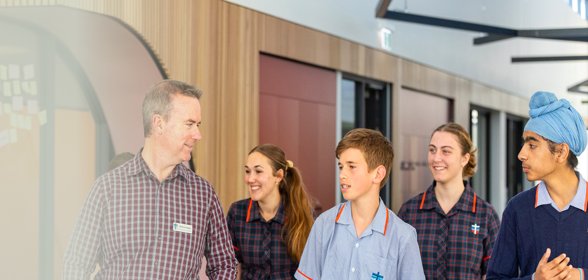 A man in a checkered shirt talking with four teenagers in school uniforms, one girl wearing a headscarf, in a modern school hallway.