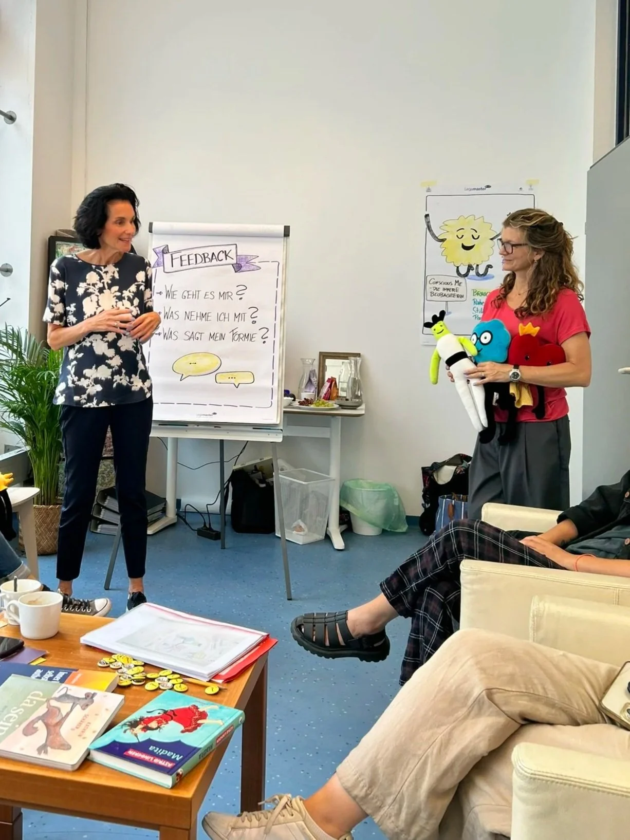 Zwei Frauen in einem Büro bei einer Präsentation. Eine steht vor einem Flipchart mit Fragen, die andere hält Stoffpuppen in der Hand.